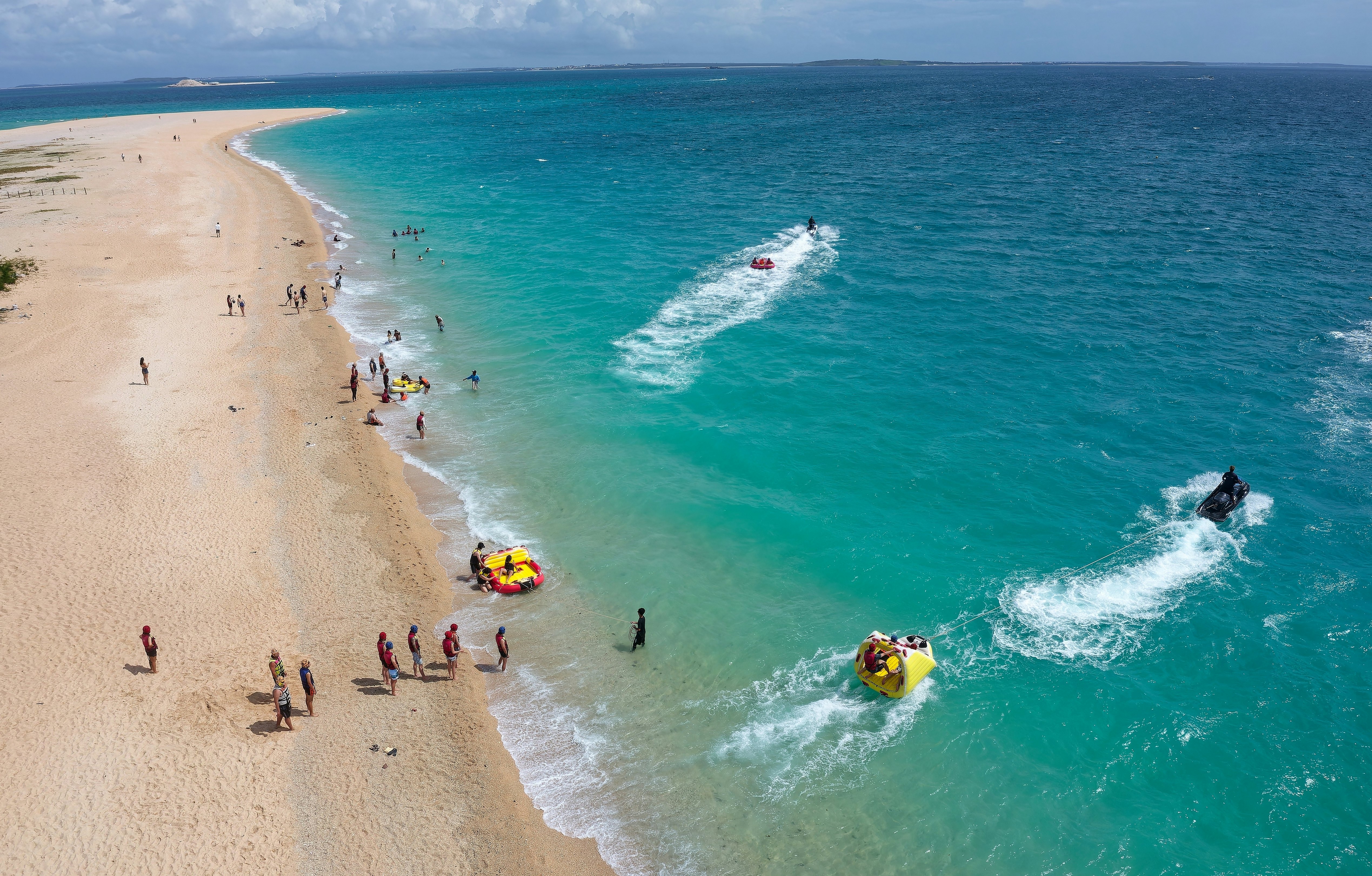 An aerial view of people on a tropical beach, with some people getting on floats pulled by boats.
