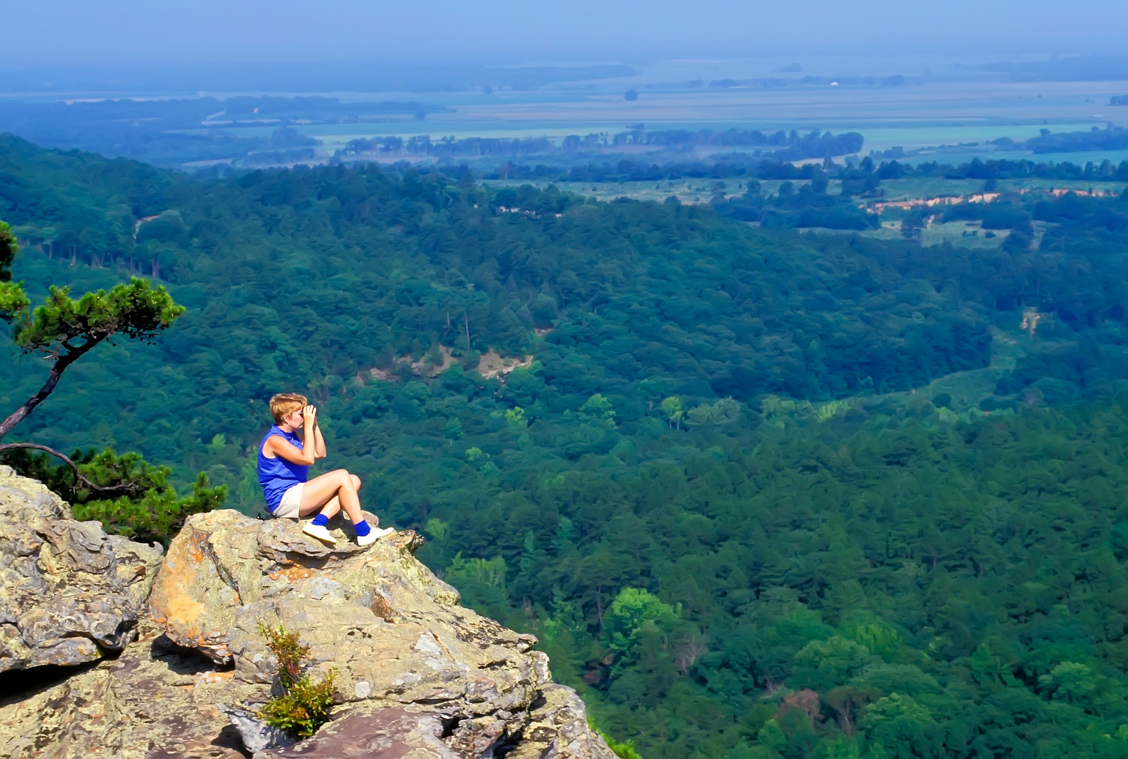 A woman looks through binoculars on a rocky outcrop overlooking a lush valley below.