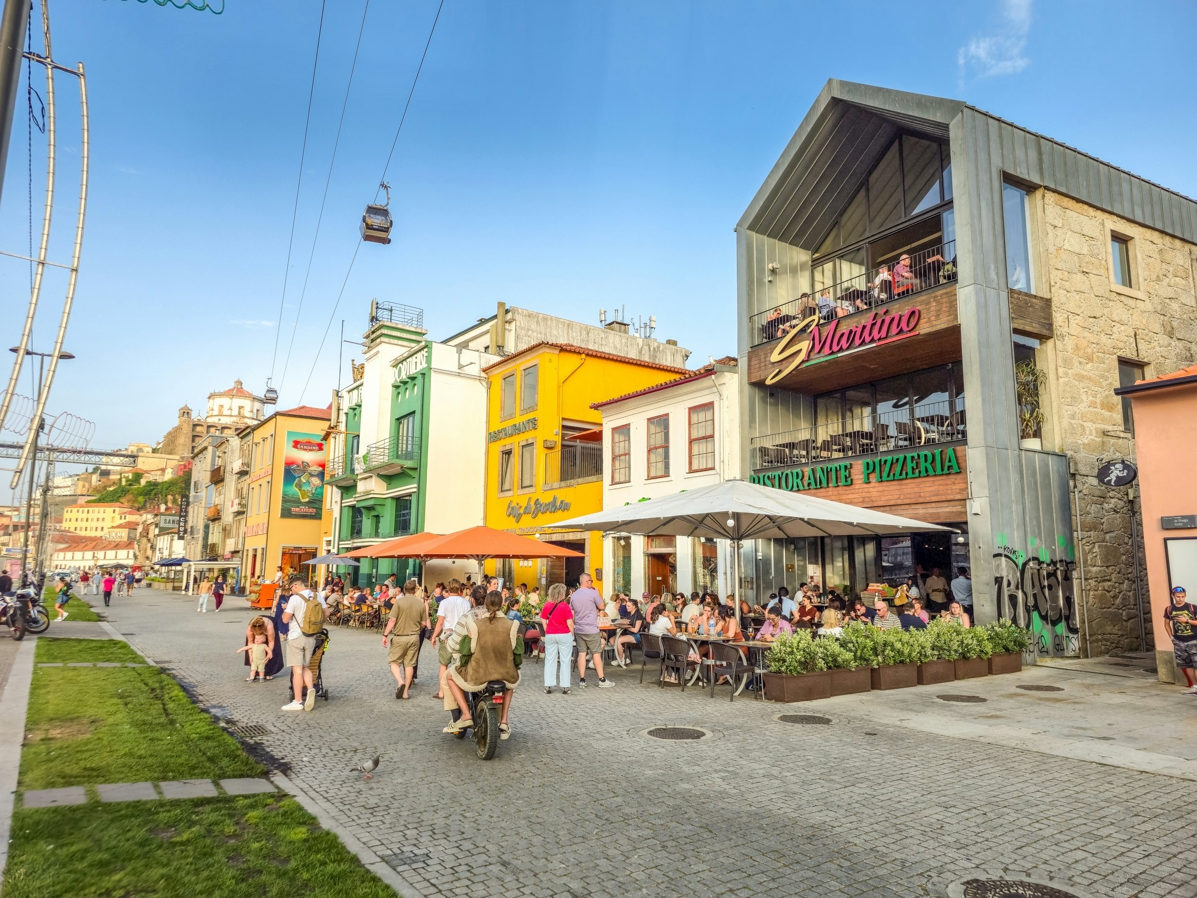 People walk along a cobbled street in front of restaurant terraces. A cable car passes overhead.