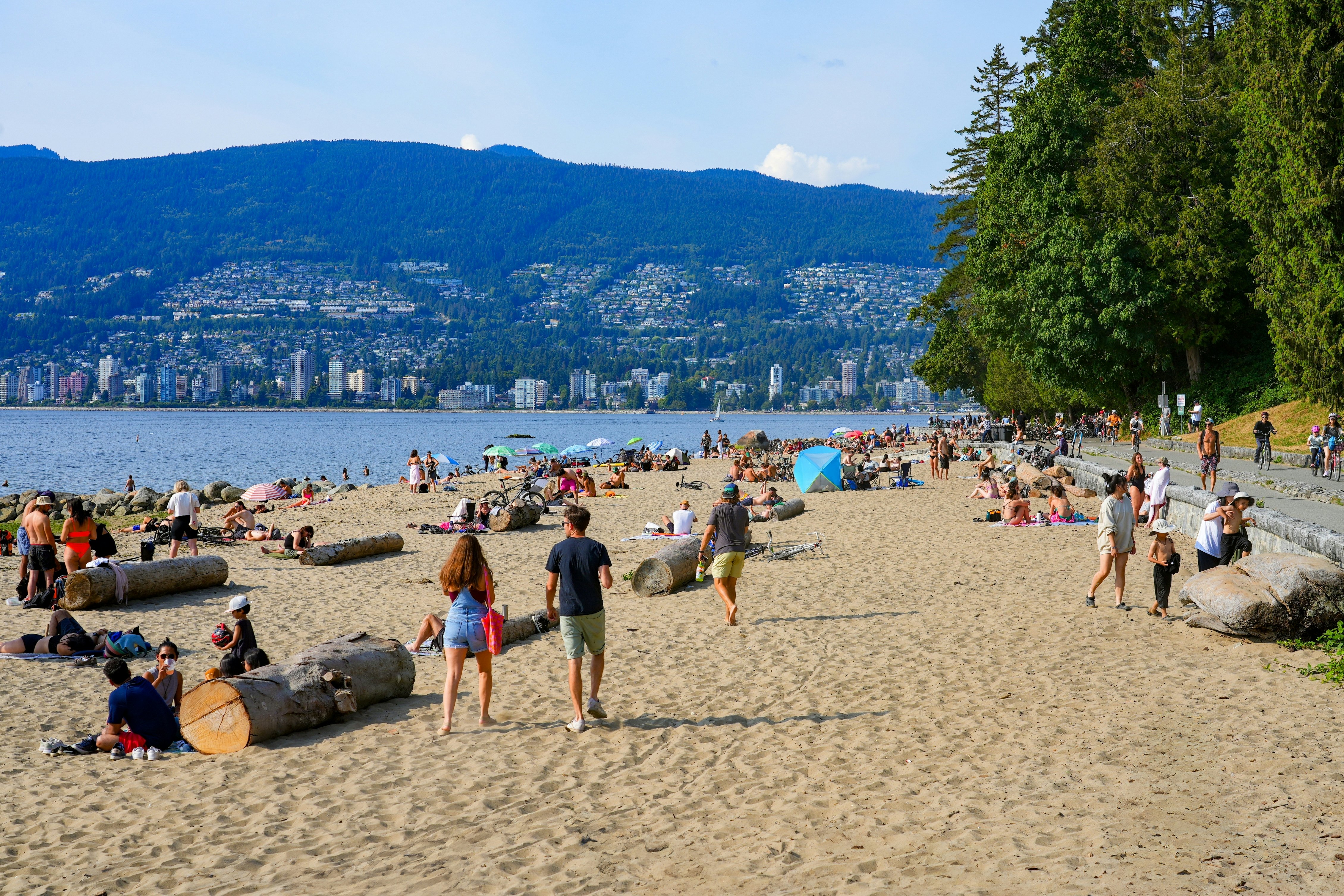 People walk and lie out on a wide beach in a city park. Towers and houses in hills are visible in the distance, across a body of water.