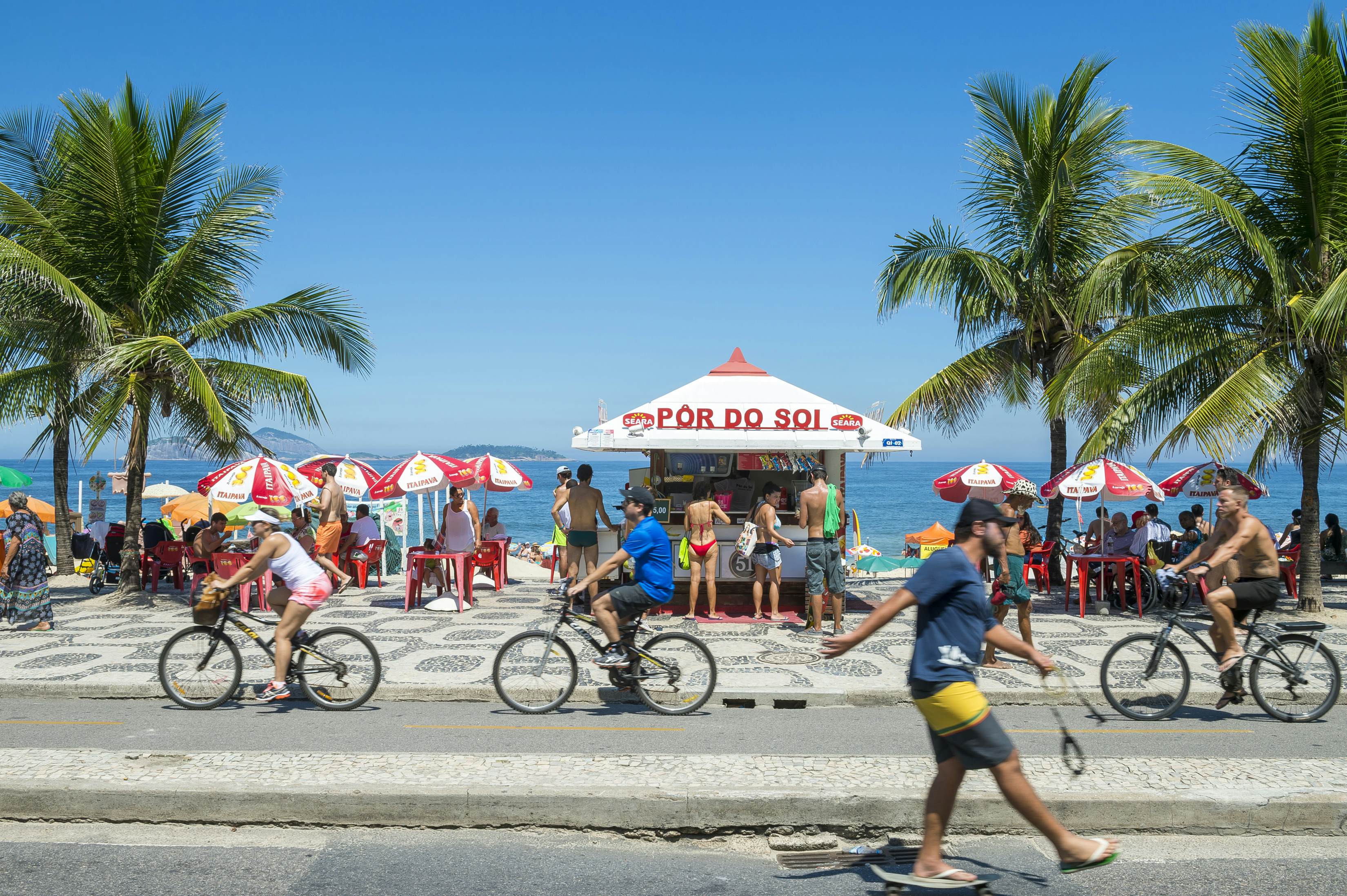  Cyclists and skateboarder pass a typical beach kiosk on the boardwalk at Ipanema Beach  