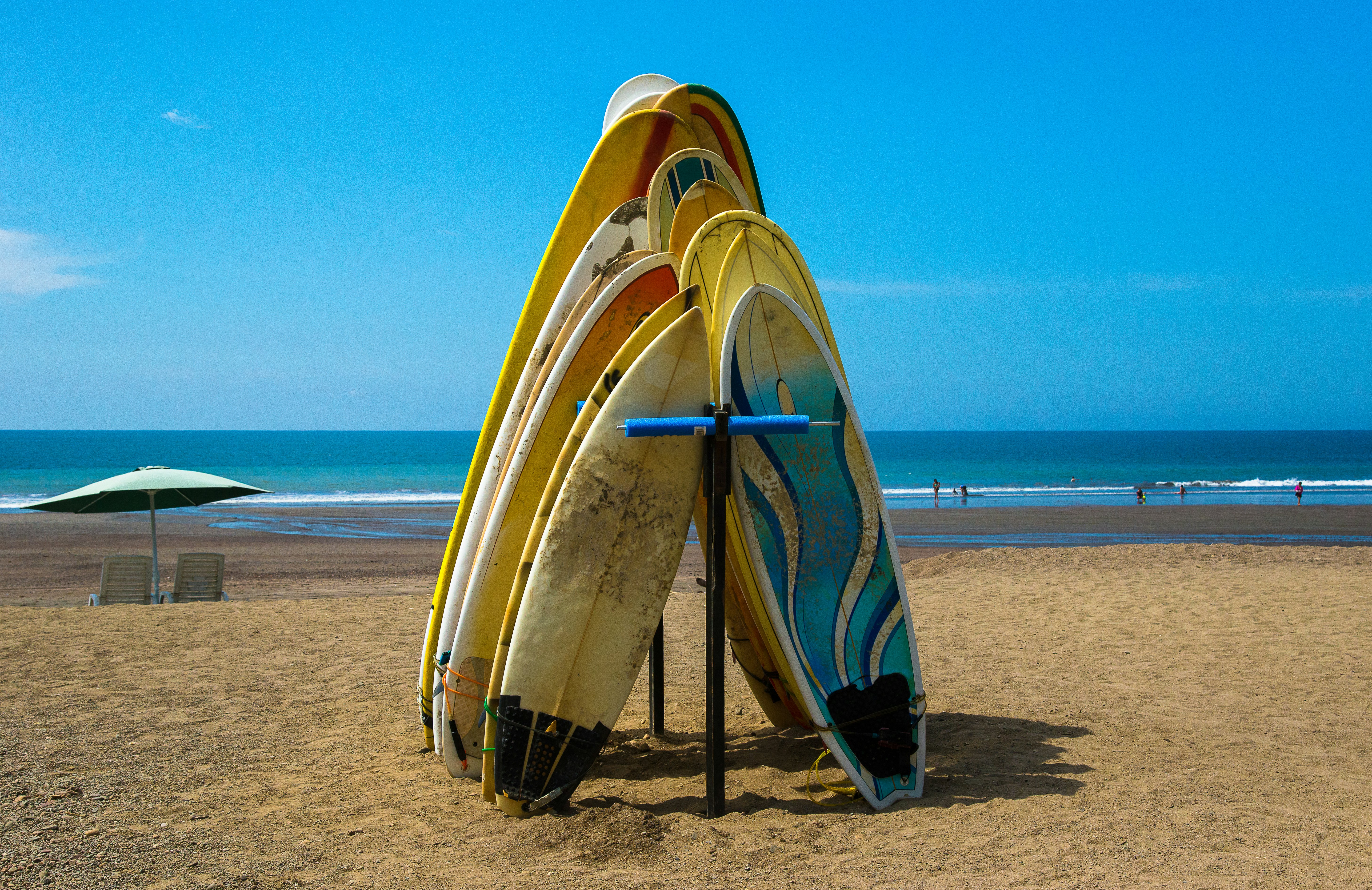 Surfboards lean against a rack on a golden sand beach under clear blue sky in Costa Rica; there is a blue umbrella with two chairs underneath by the blue water.