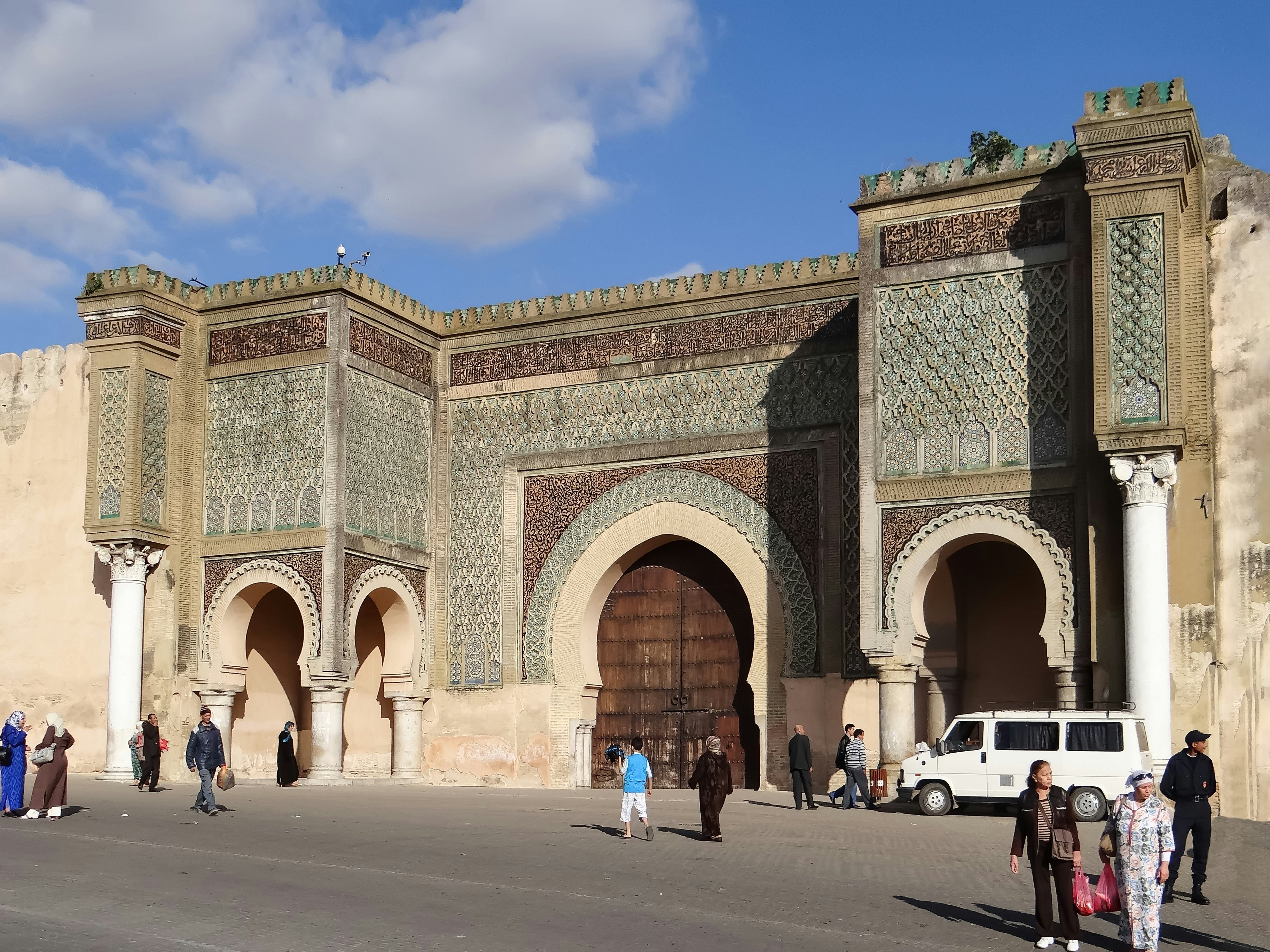 People walking near an elaborate city gate in Morocco; it has large archways, columns, wooden doors and decorated tiles and carvings.