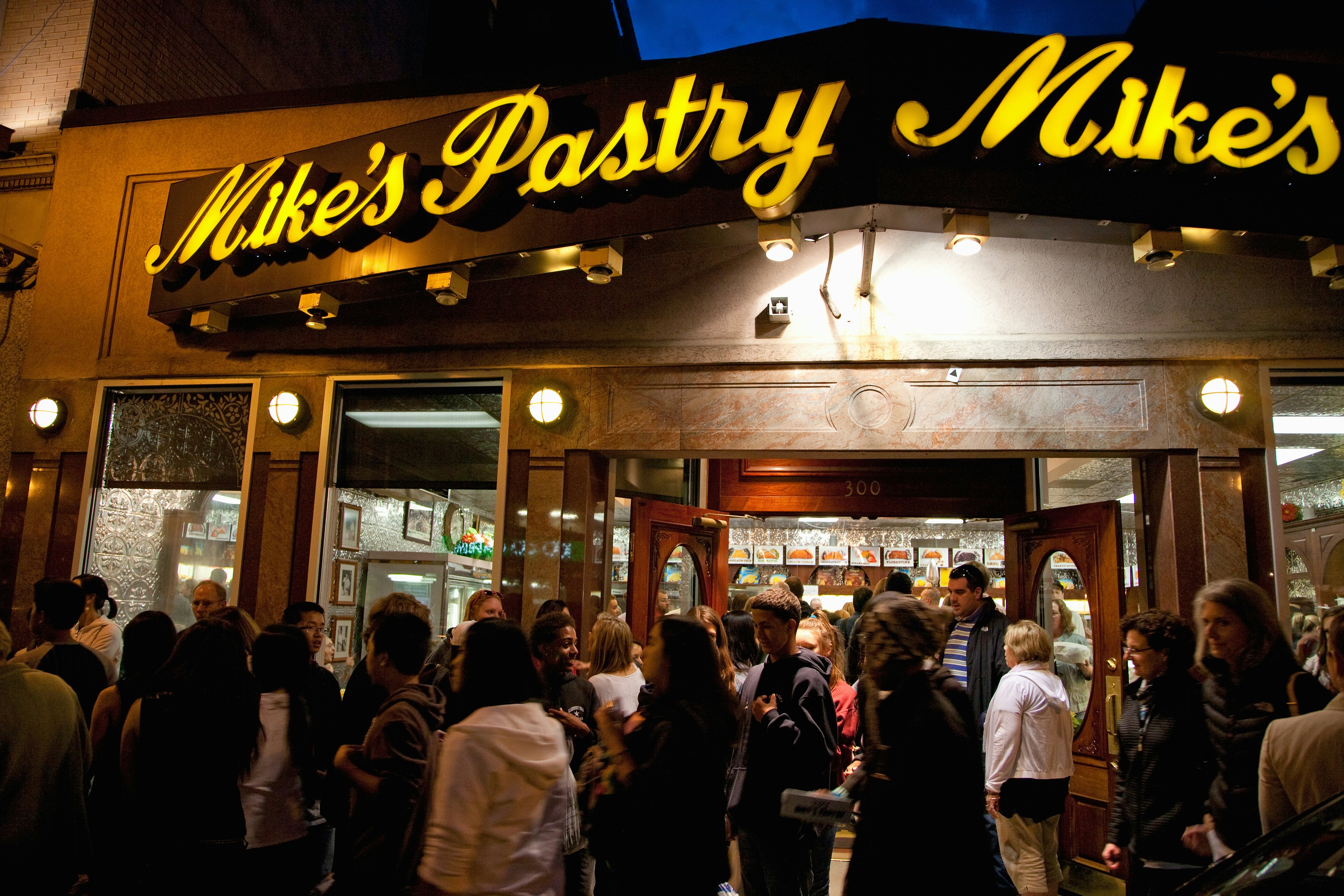 A crowd of people stand in front of a restaurant with illuminated signage at night.