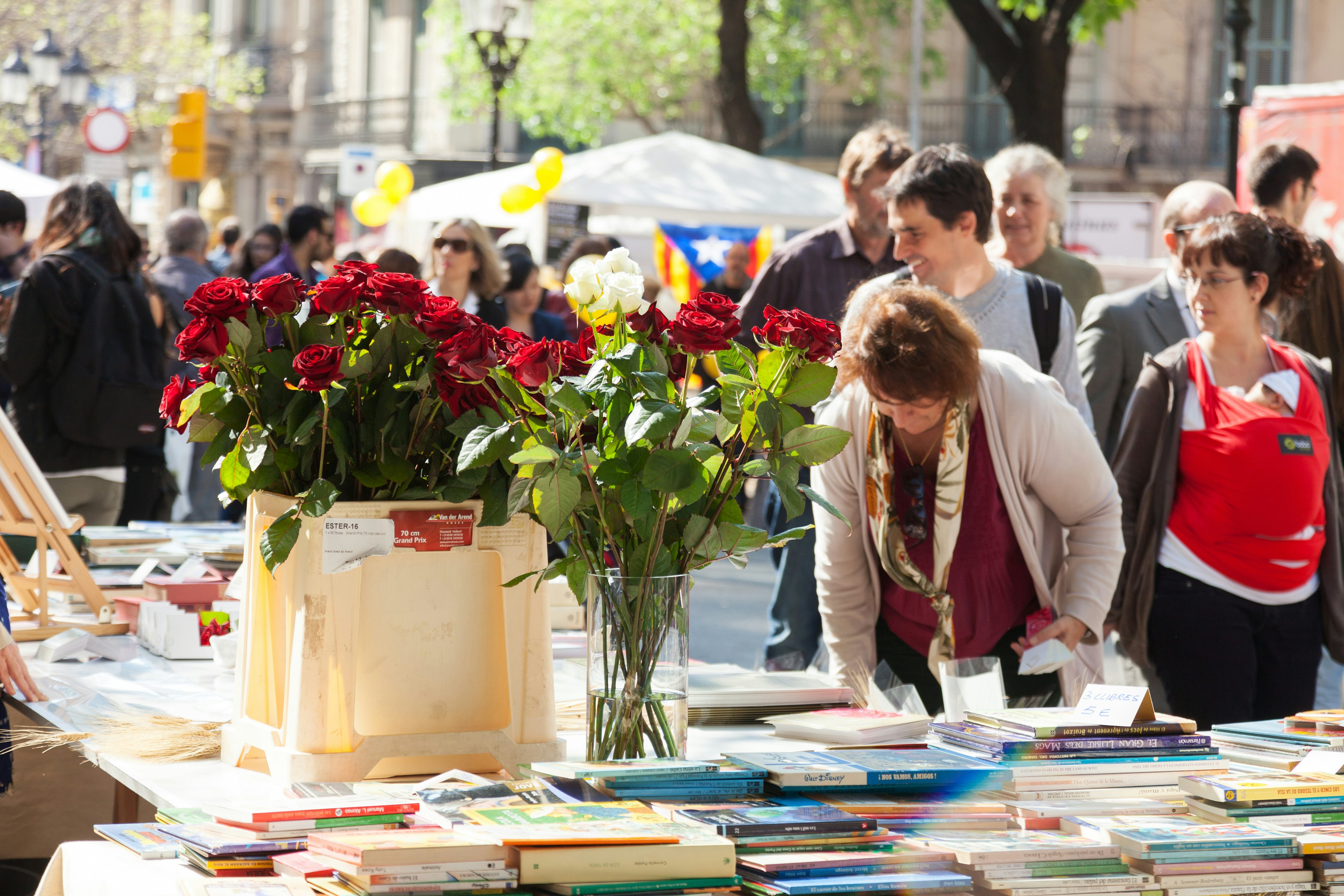 People browse book stalls at an open-air market on a sunny day. Roses are placed in vases on the tables.