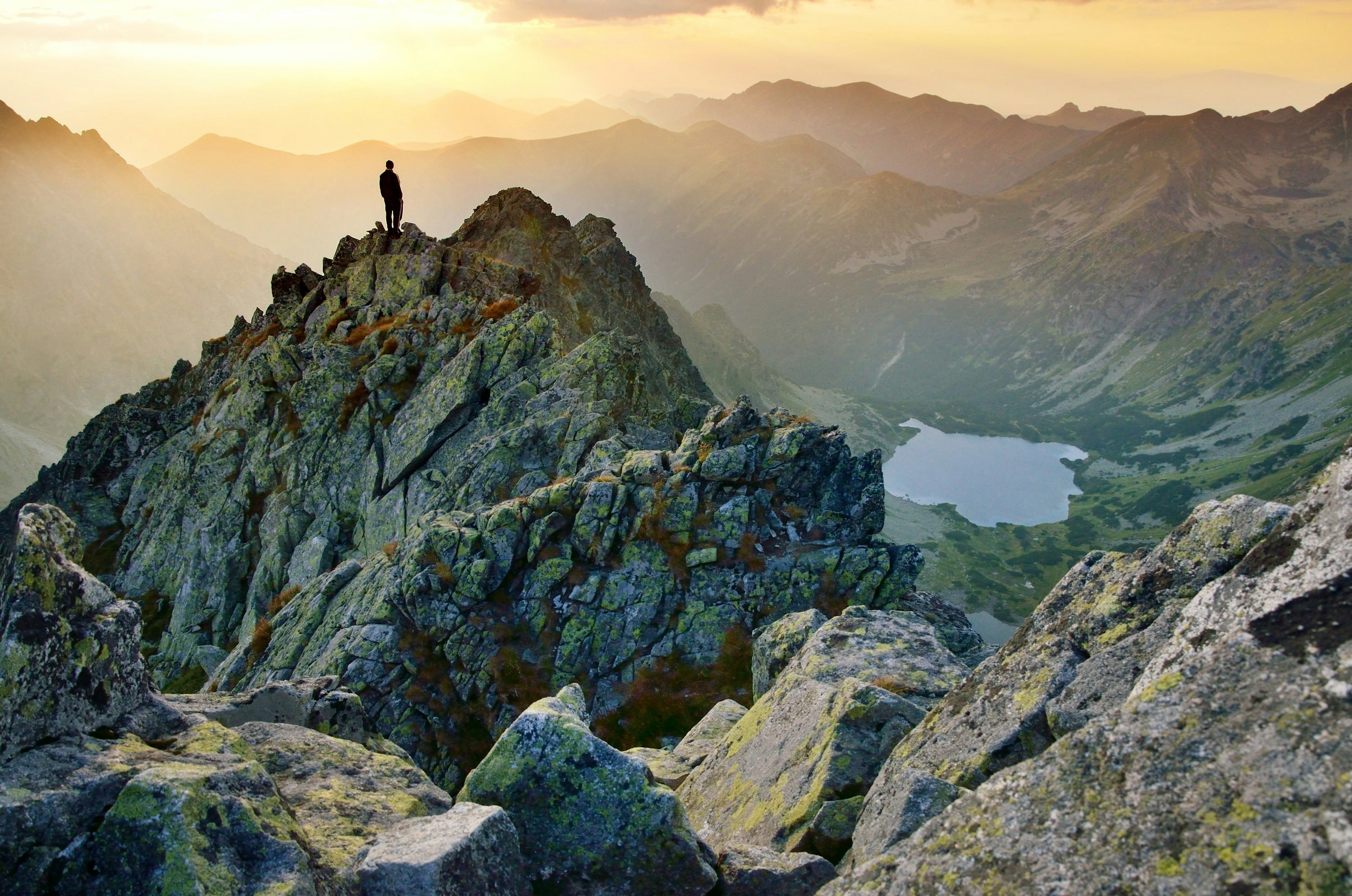 A single hiker stands on a rocky peak in Slovakia, looking over a mountain range with a lake far below.
