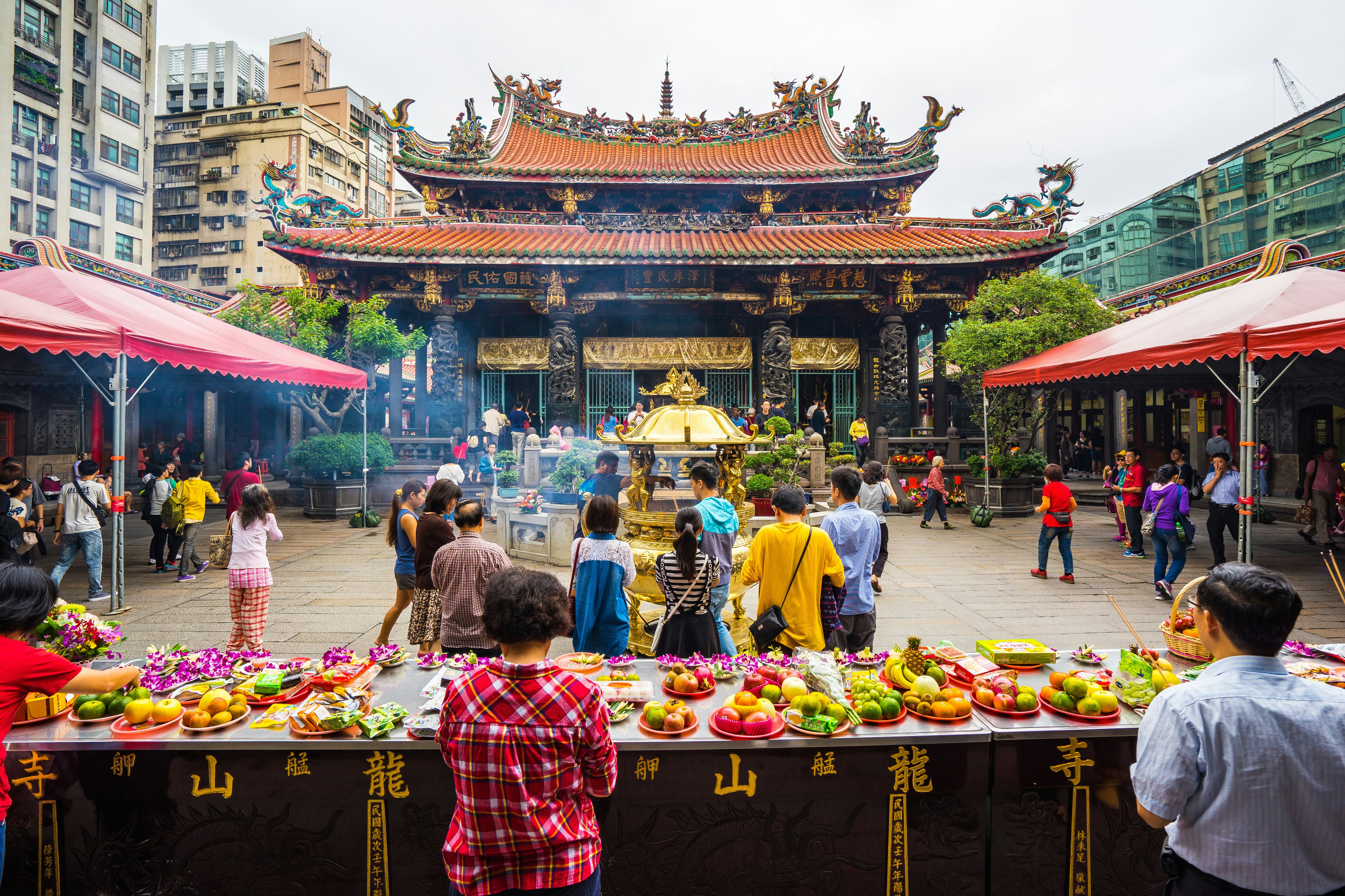 People stand in front of table with offerings of fruit at a temple. The elaborately roofed sanctuary is visible across a courtyard.