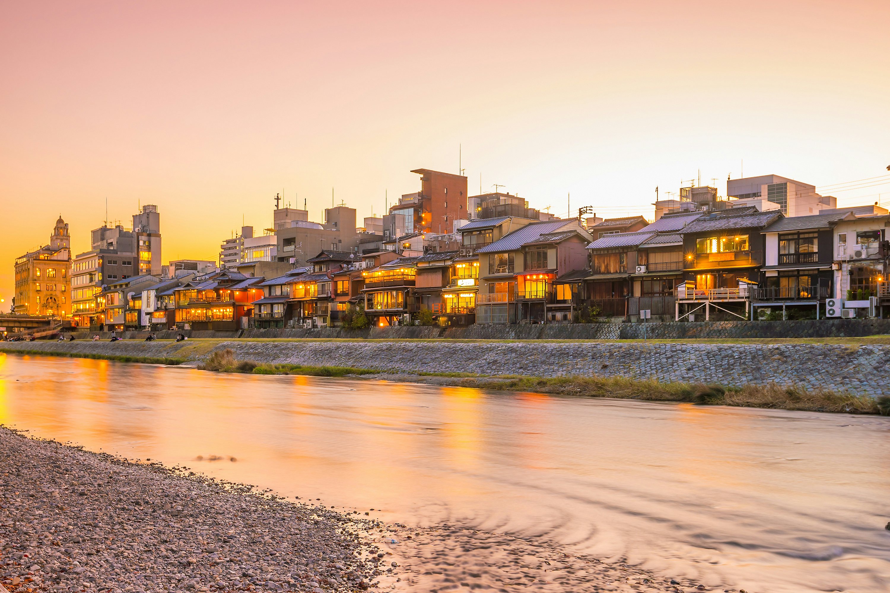 Traditional Japanese wooden houses on the banks of a river as the sunset casts an orange glow across the sky.