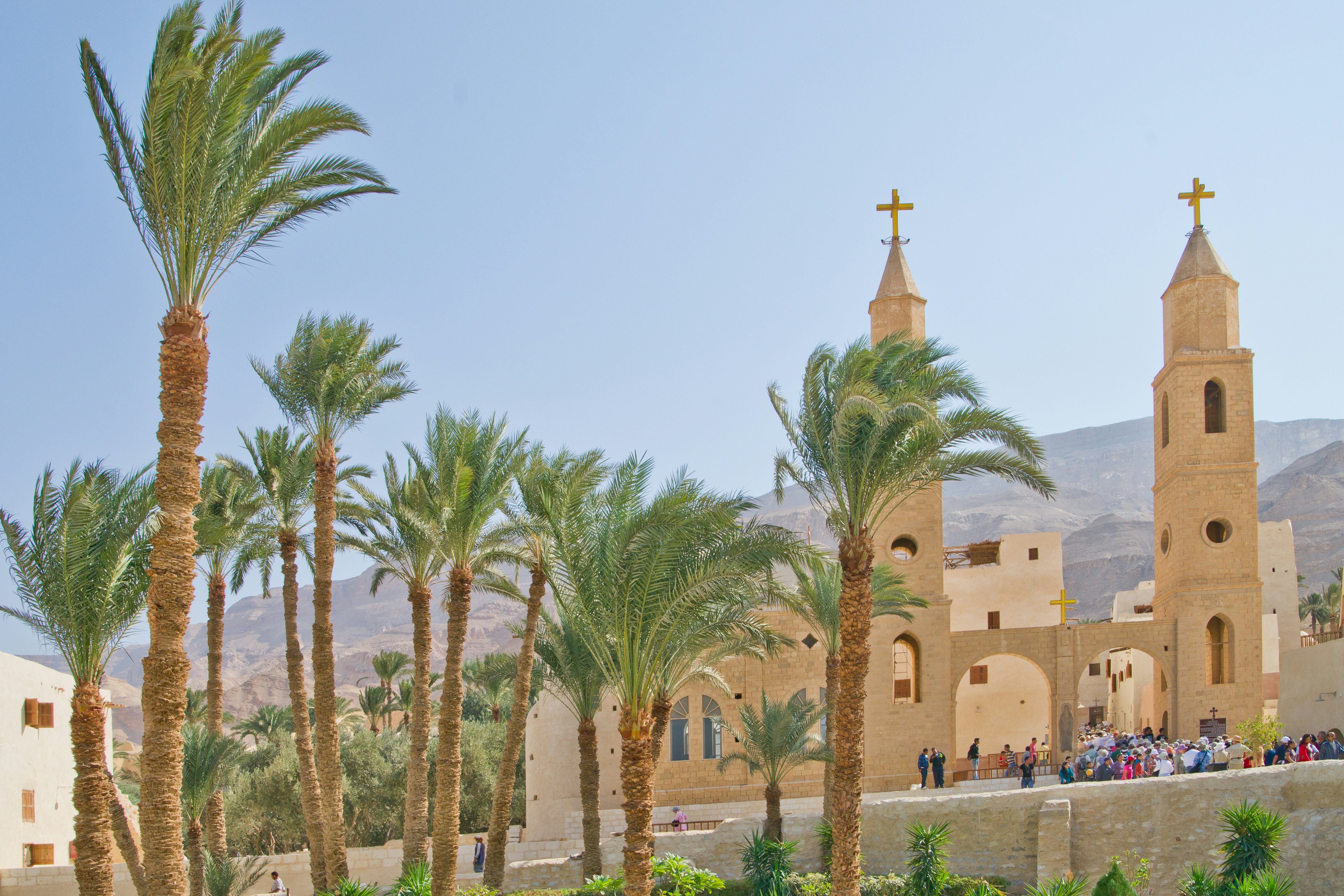 Pilgrims enter a monastery surrounded by palm trees in a hilly desert area.