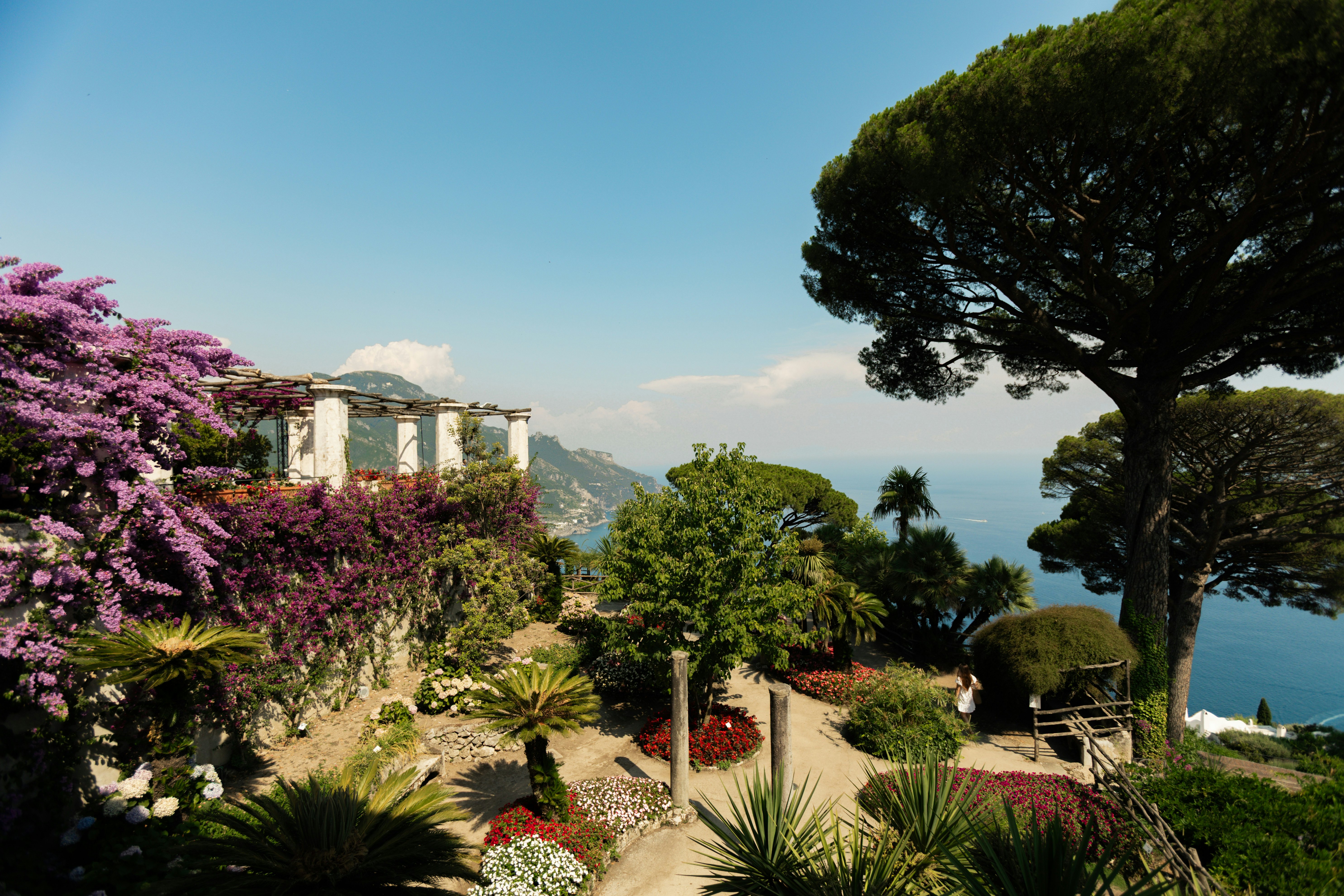 A large formal garden and patio perched on a hillside overlooking the sea and forest-covered mountains in the distance on a sunny day.