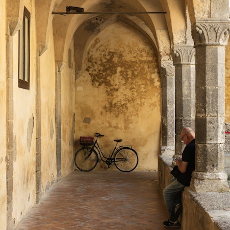 A man leans along a column in a stone courtyard with a parked bicycle in the far distance.
