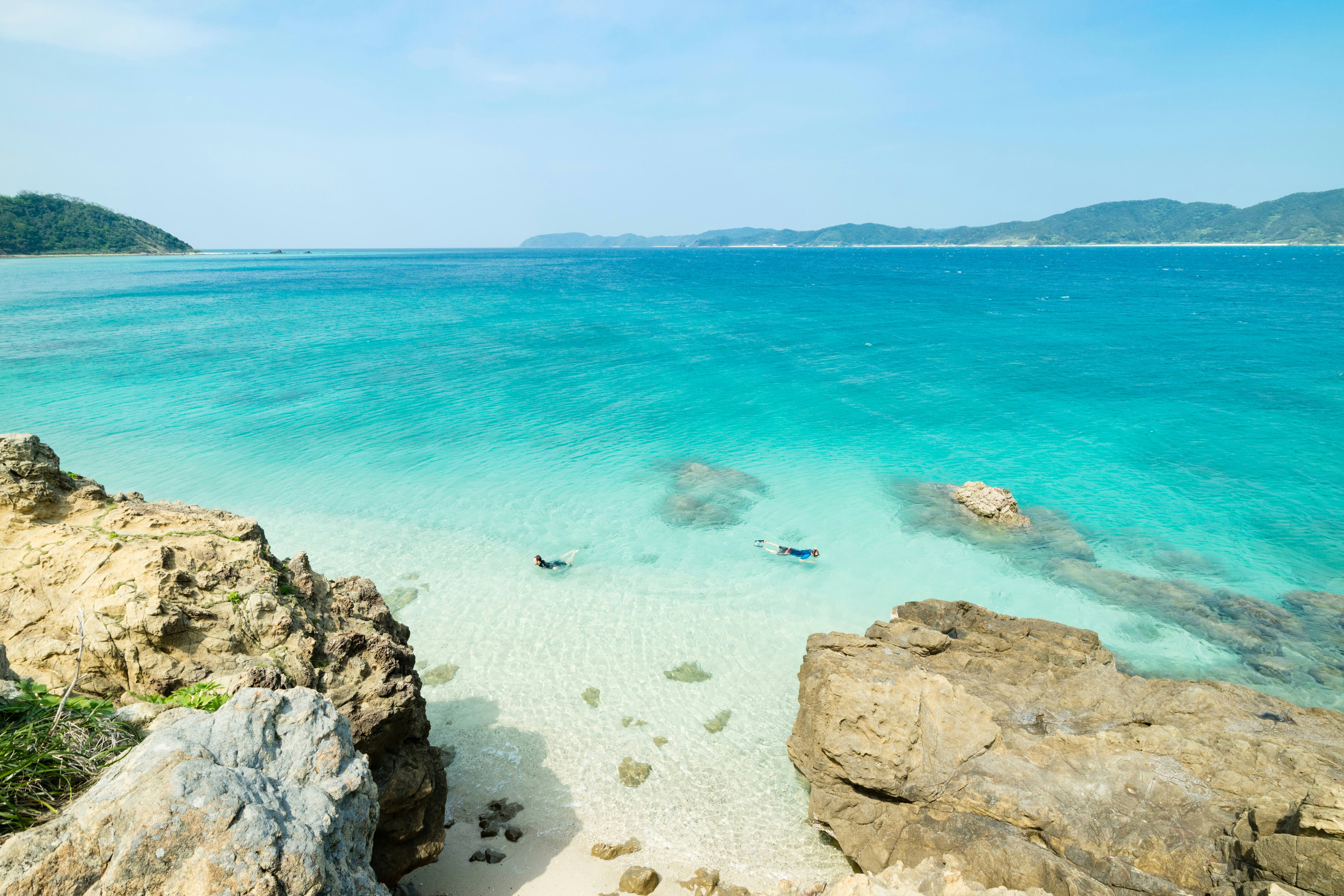 Two snorkelers explore crystal-clear ocean near a rocky shoreline.