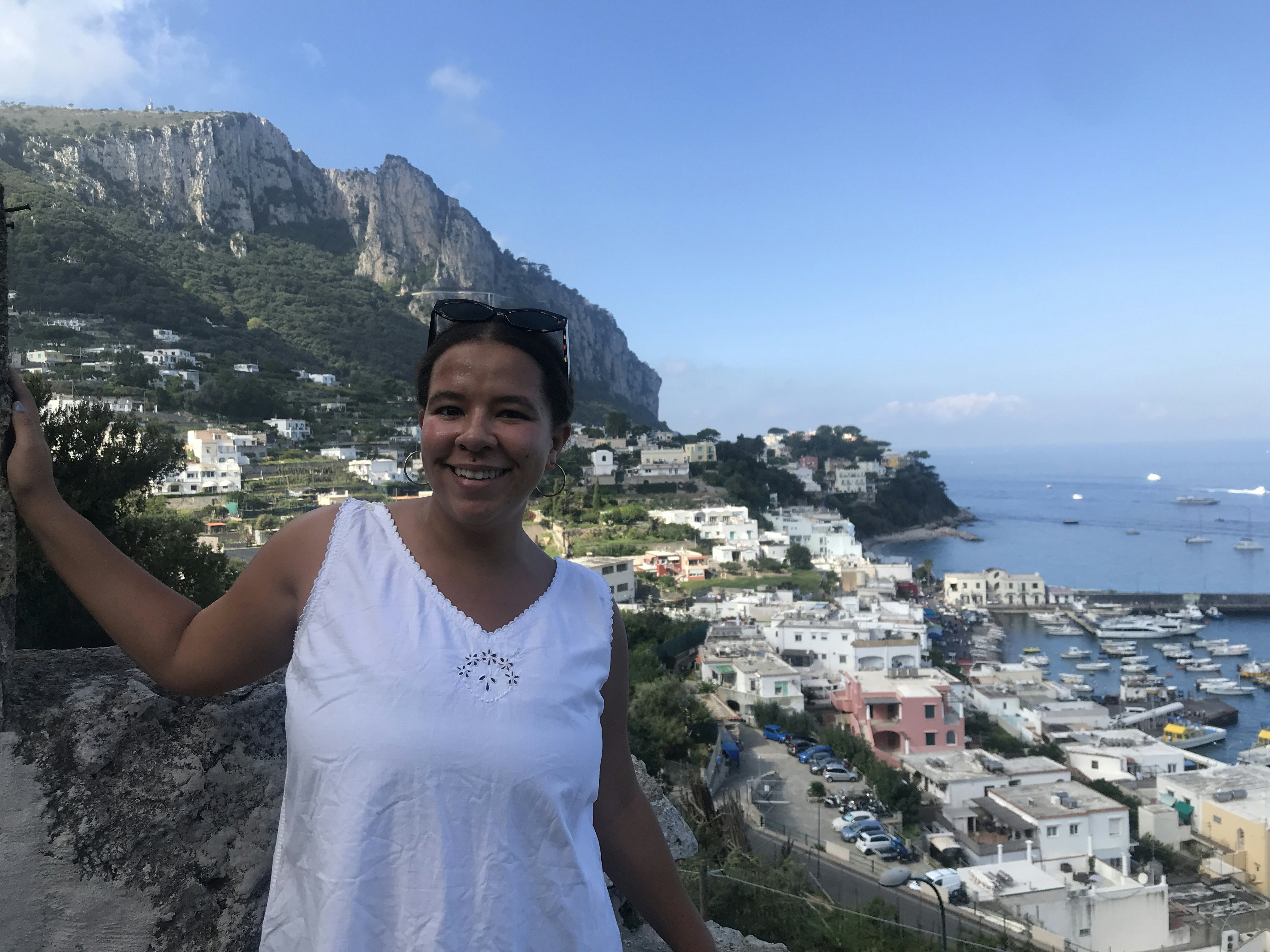 A woman standing on a cliff above a town and ocean on a sunny day.