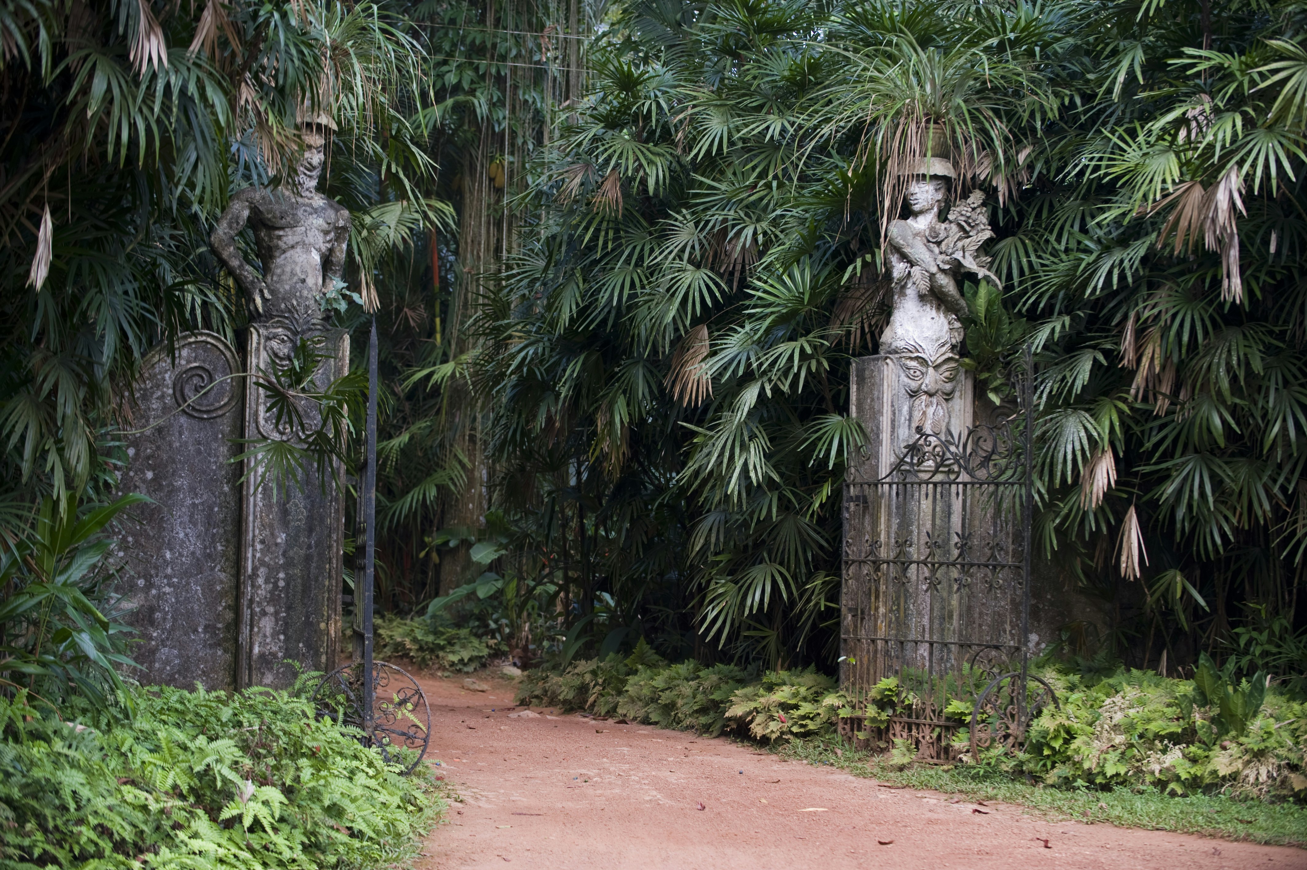 A dirt road flanked by two sculptures on posts with an open gate between them. Greenery is on both sides of the road.