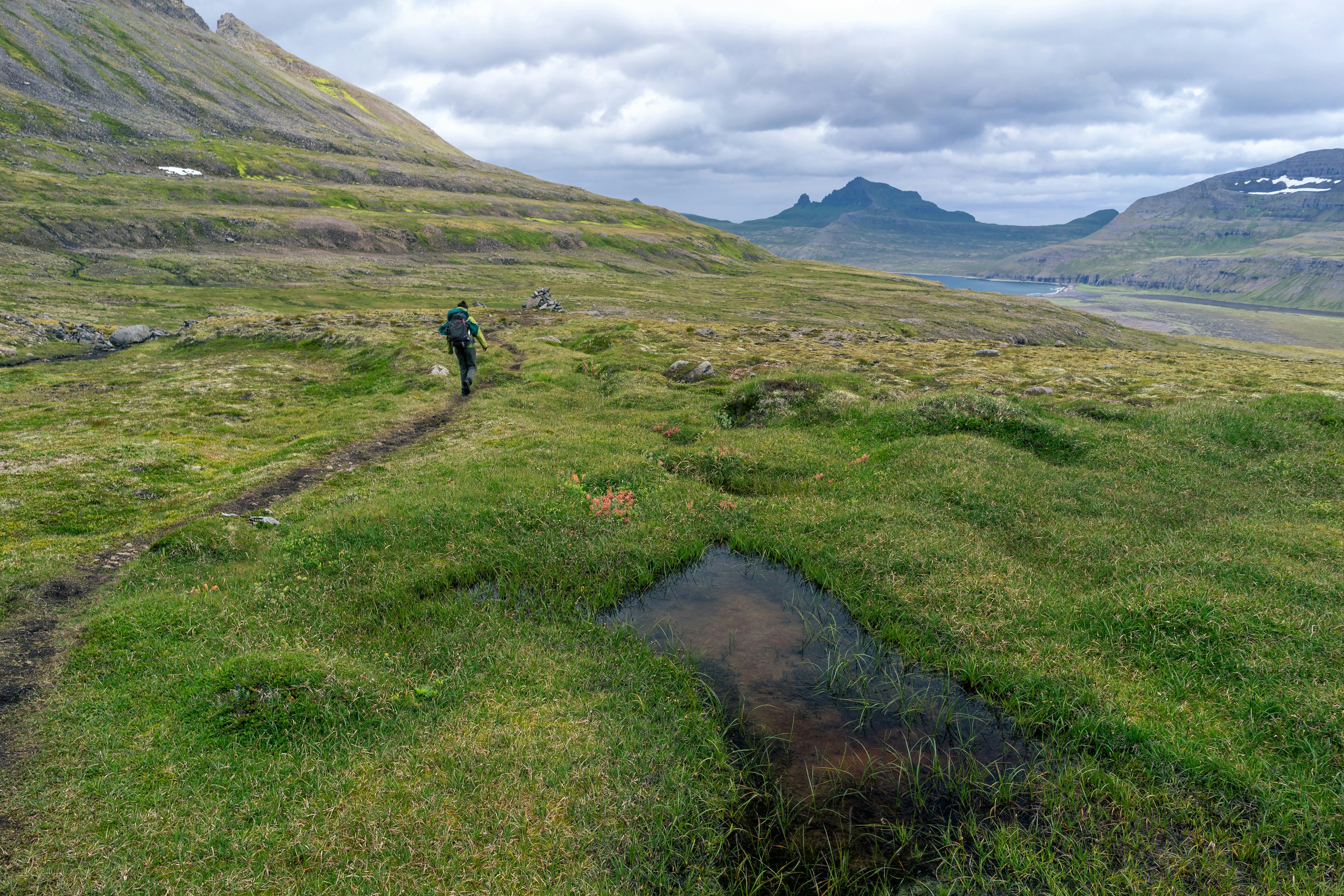 A hiker walking on a narrow dirt trail through grassland in Iceland, with a mountain peak in the distance, under cloudy skies.