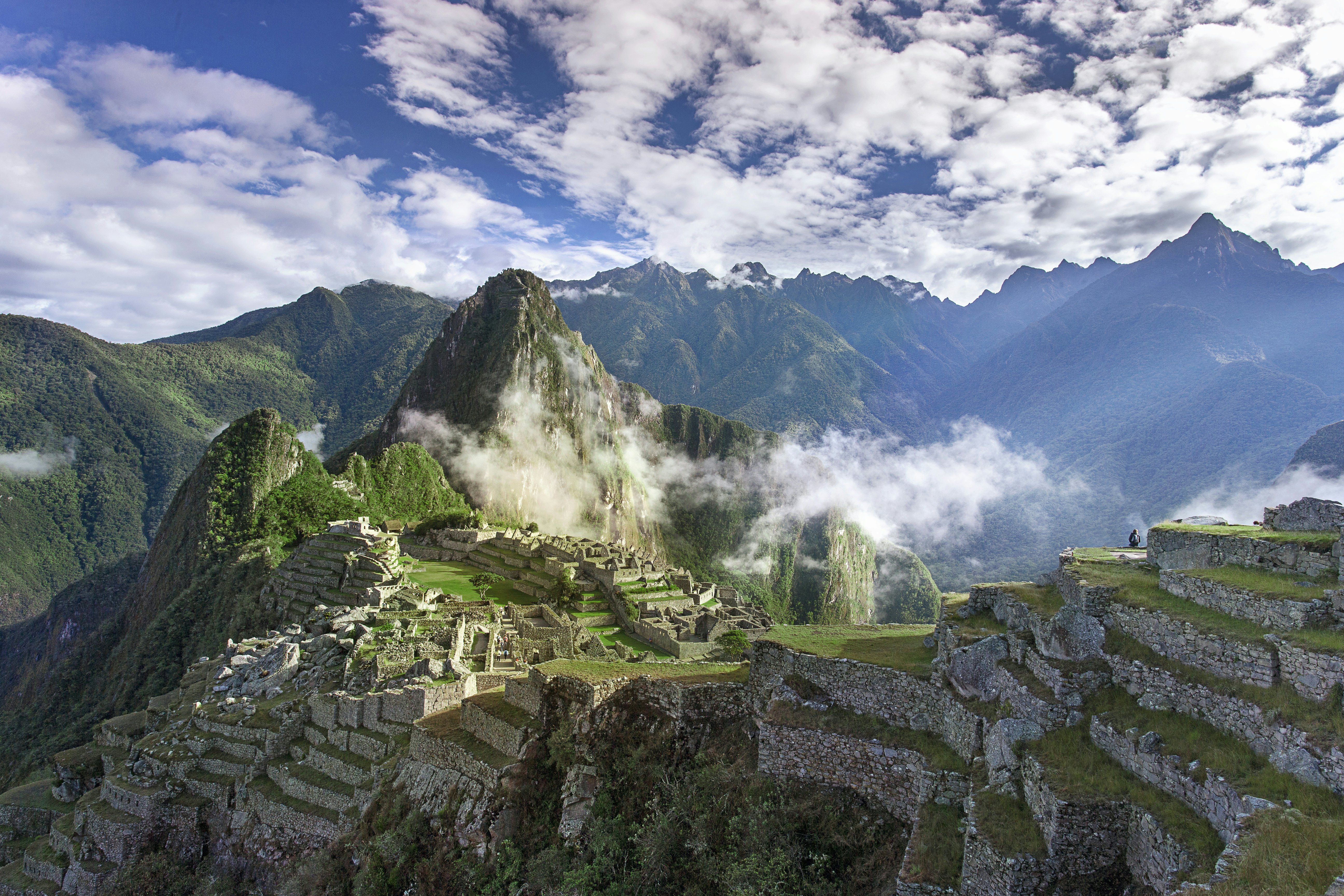 Ancient city walls built into tiers on steep rocky outcrops at high altitude, as wisps of cloud float over.