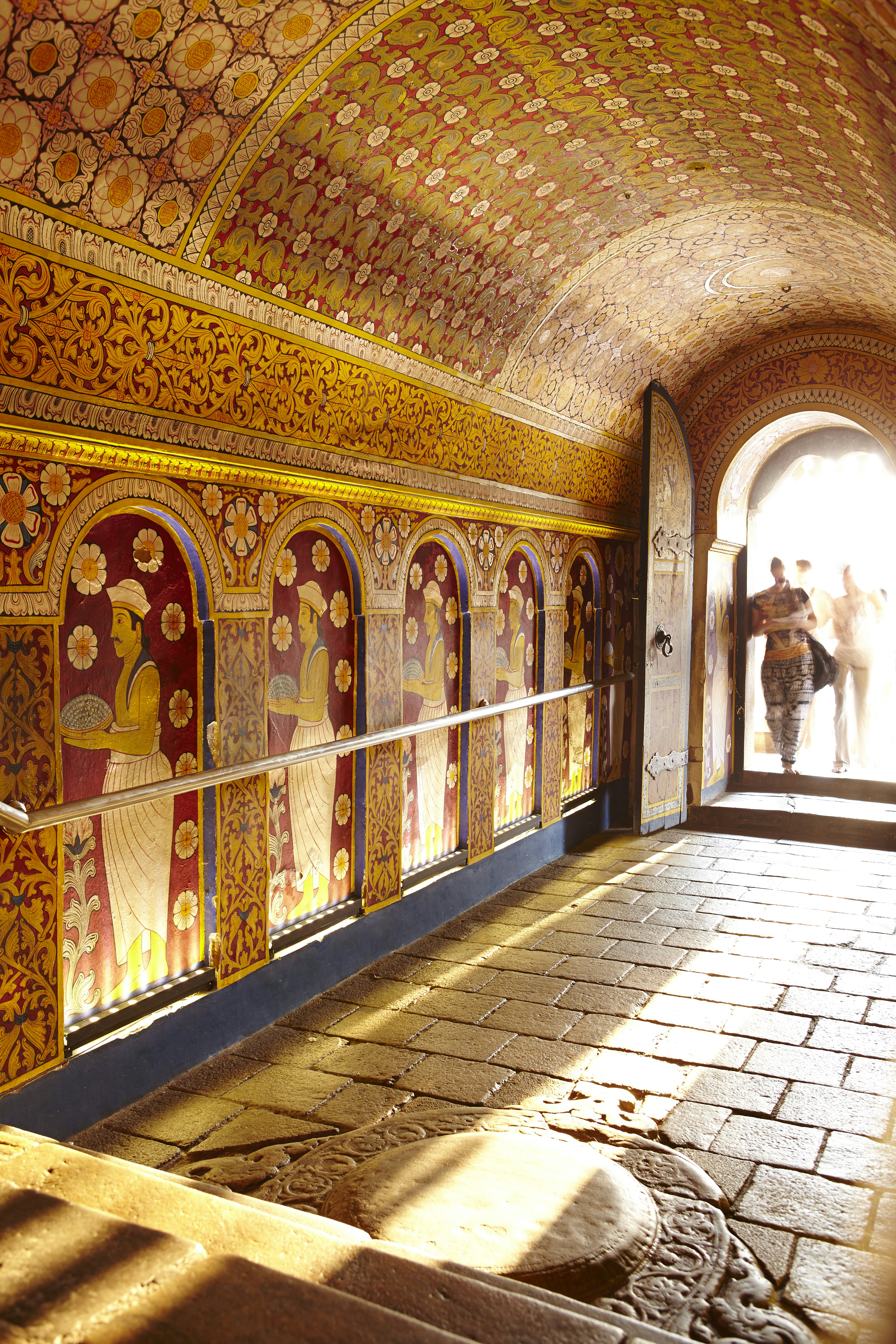 Sun shines into a room with an intricately decorated wall and stone flooring. People are standing in the doorway.