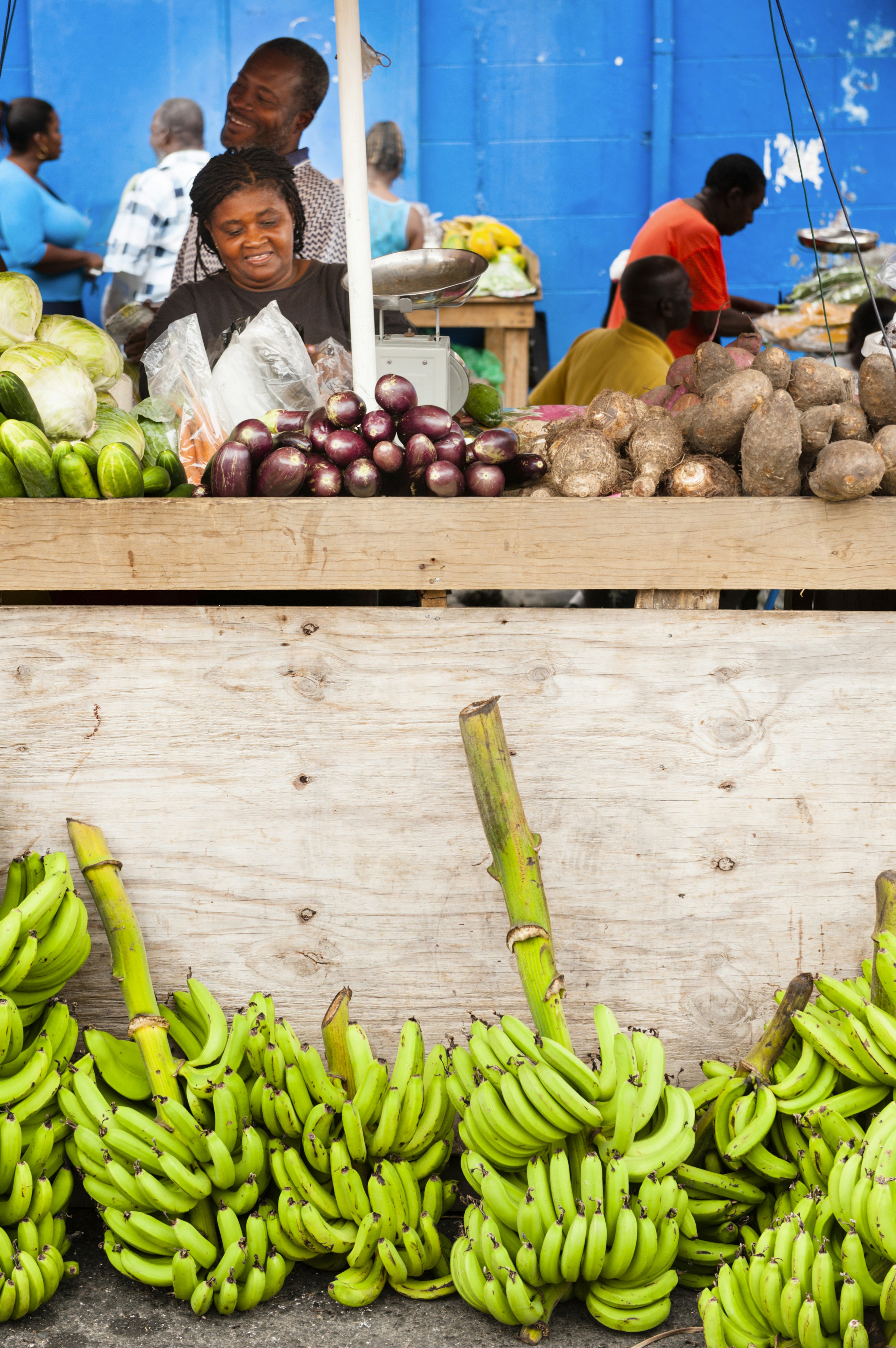 A woman oversees a food stall at a fresh produce market. There are piles of bananas at the foot of the stall.