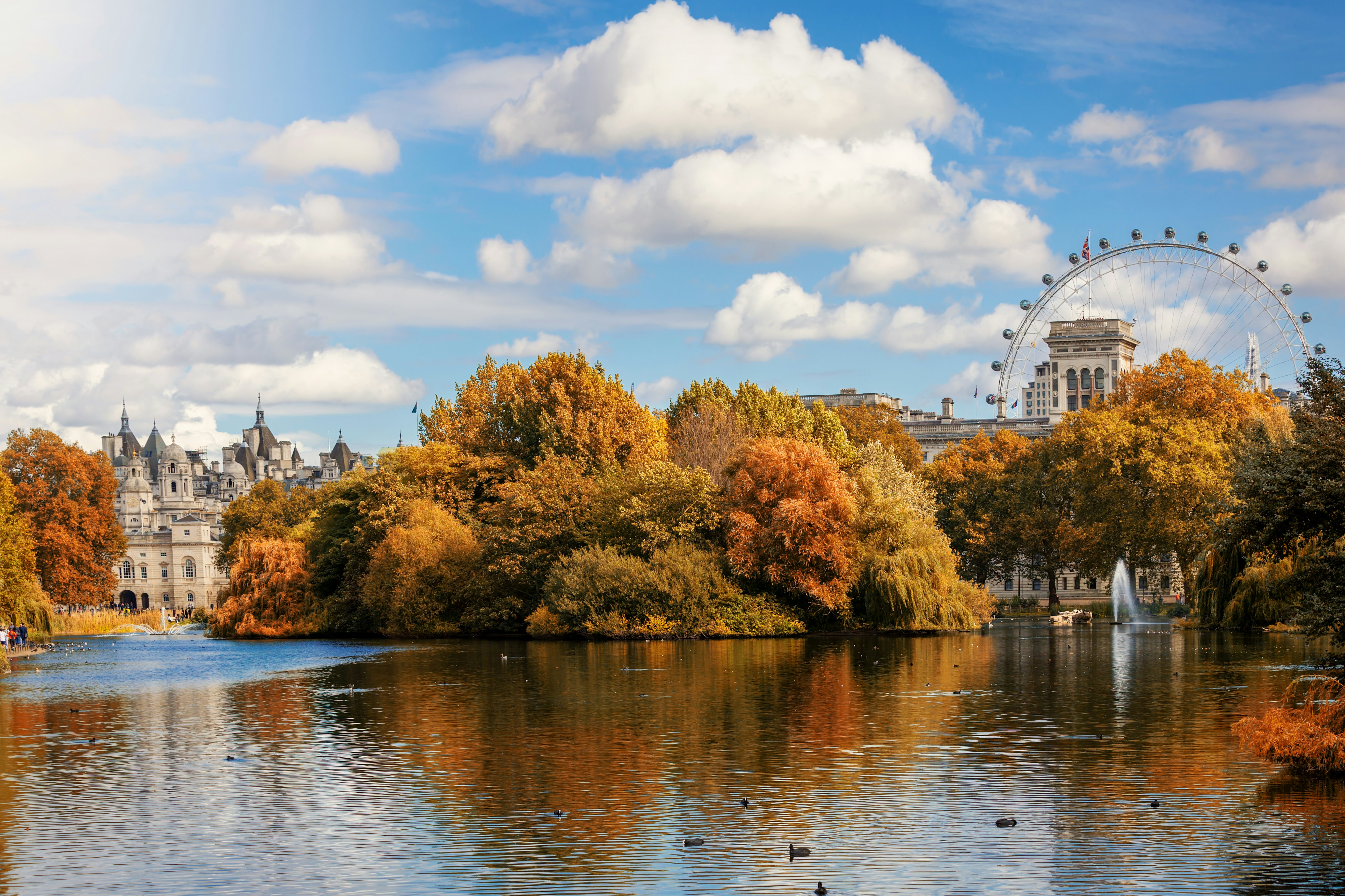 A large lake surrounded by trees with leaves that have turned golden brown in the fall. A Ferris wheel stands in the distance.