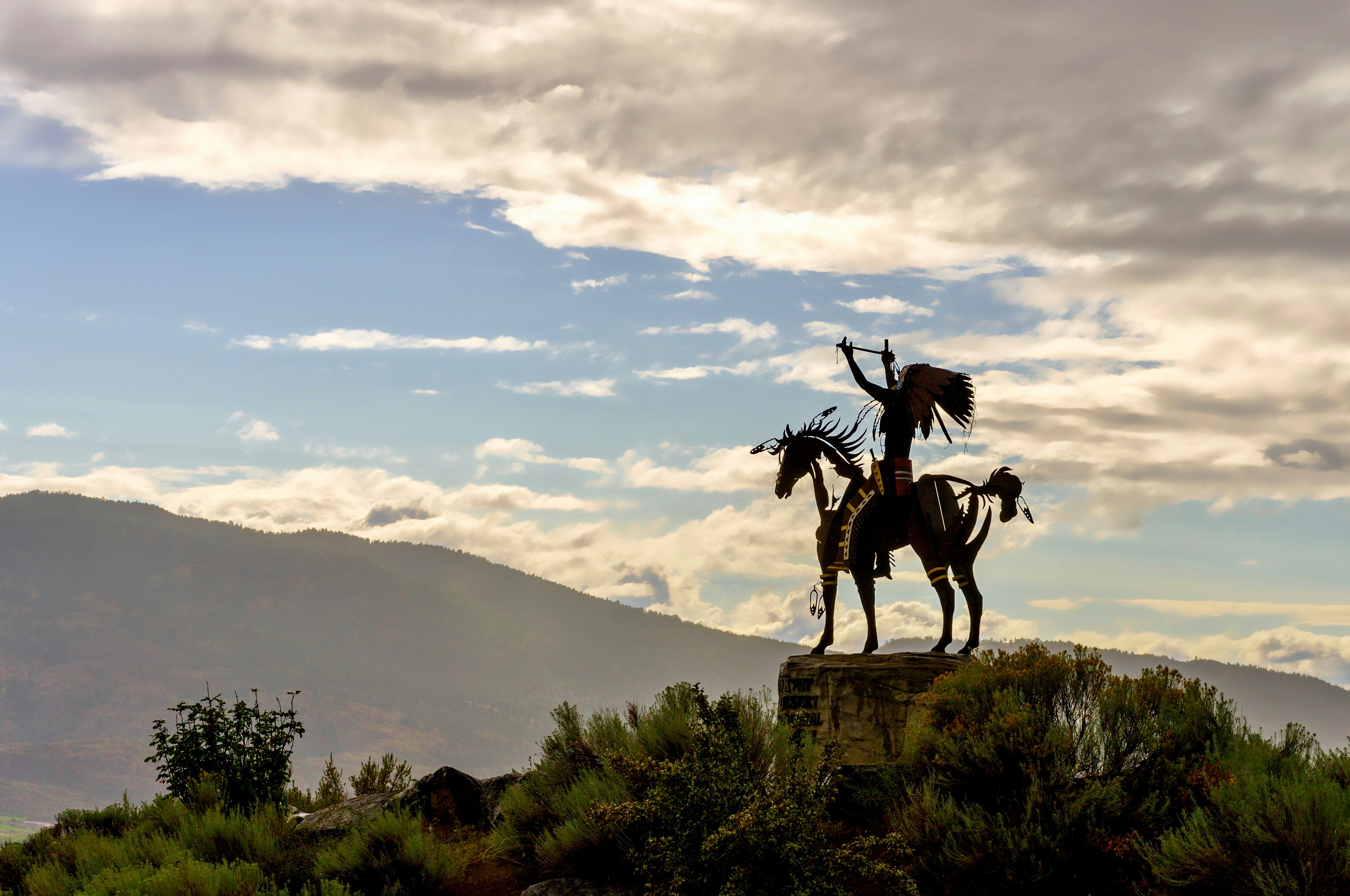 A silhouetted statue of an Indigenous warrior with moutains and clouds in the distance.