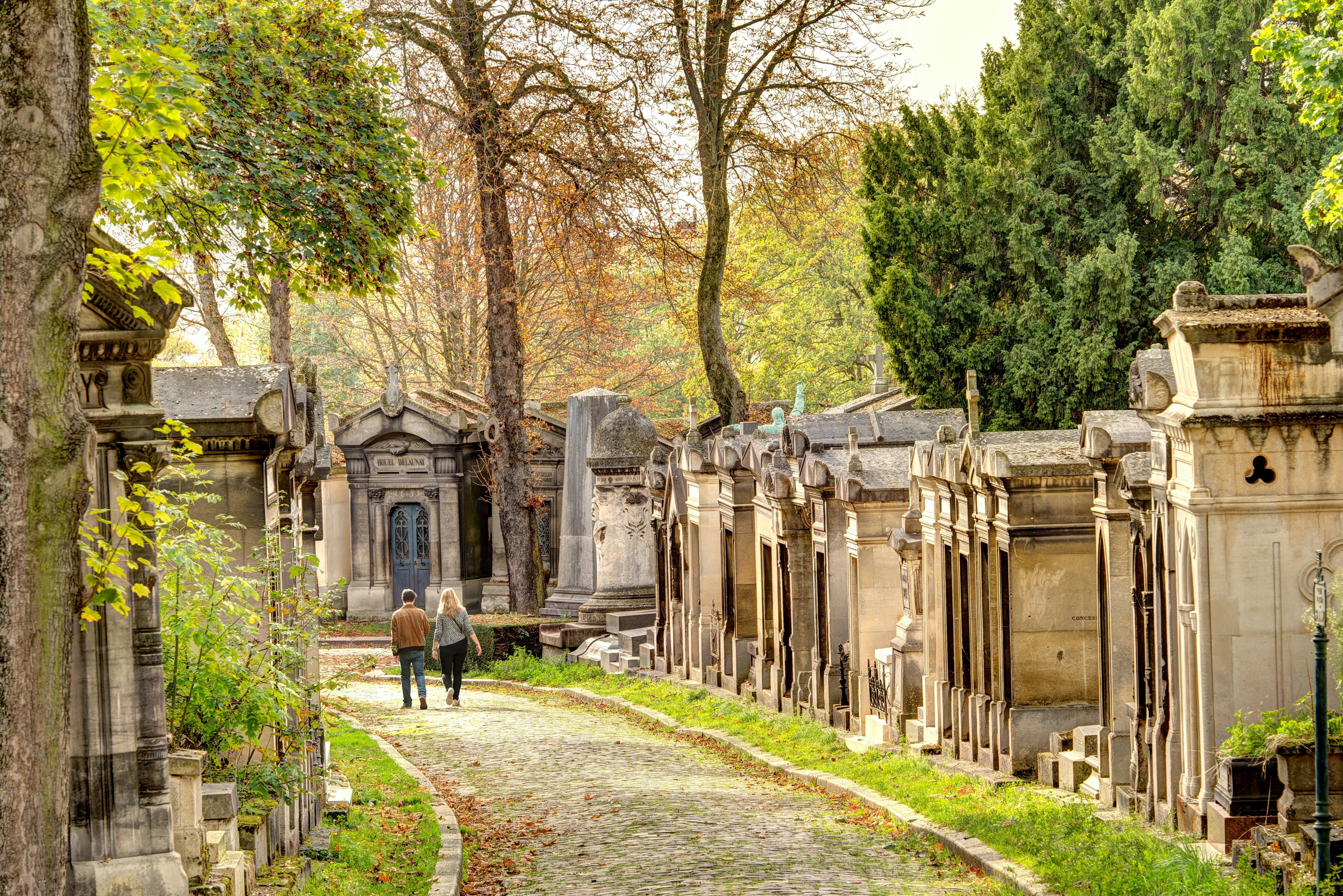 Two people walking on a cobblestone path in a cemetery with mausoleums on either side on an autumnal day.