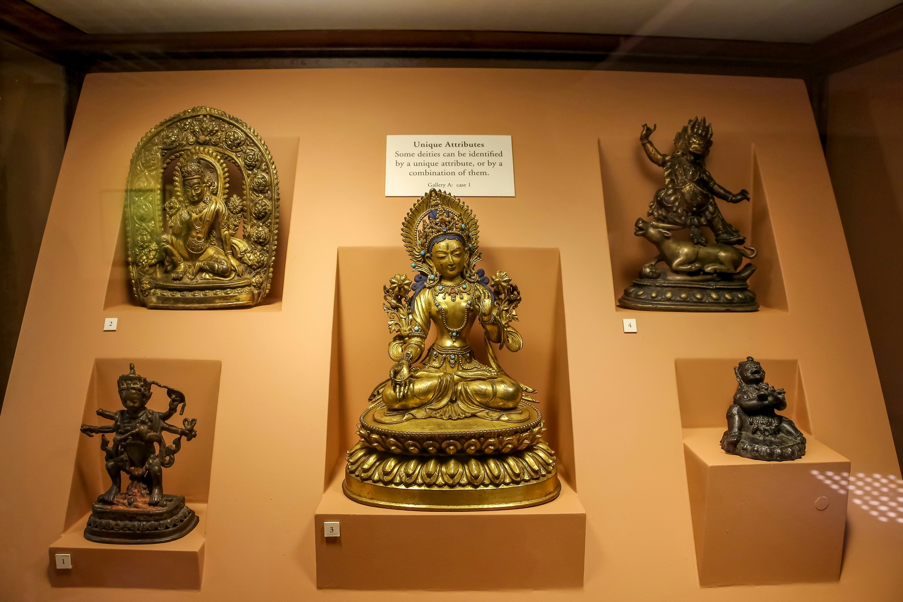 A bronze Buddha surrounded by Hindu gods figurines on orange background in a museum display