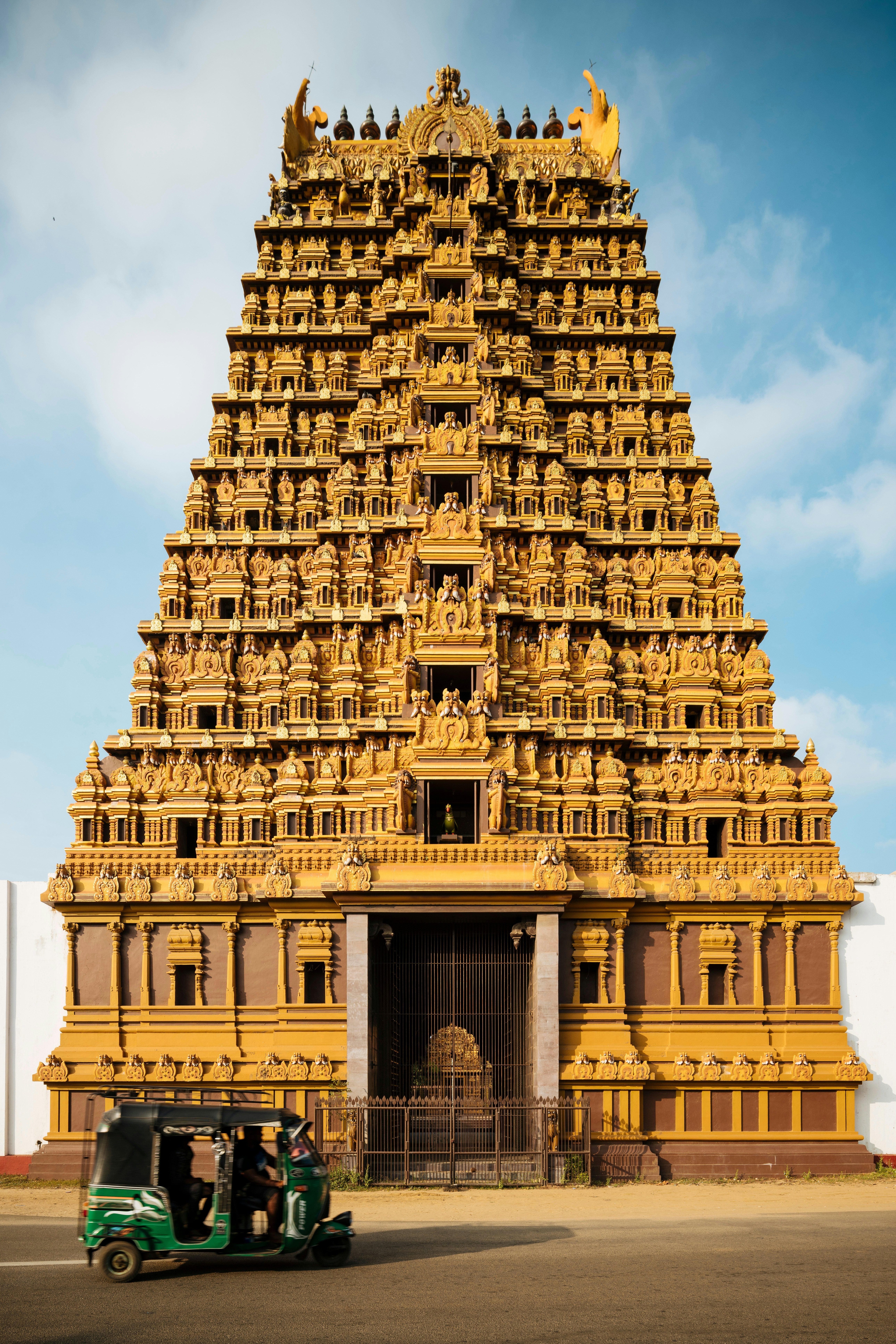 A tall, ornate gold building with a green tuk tuk driving by from the left in the foreground