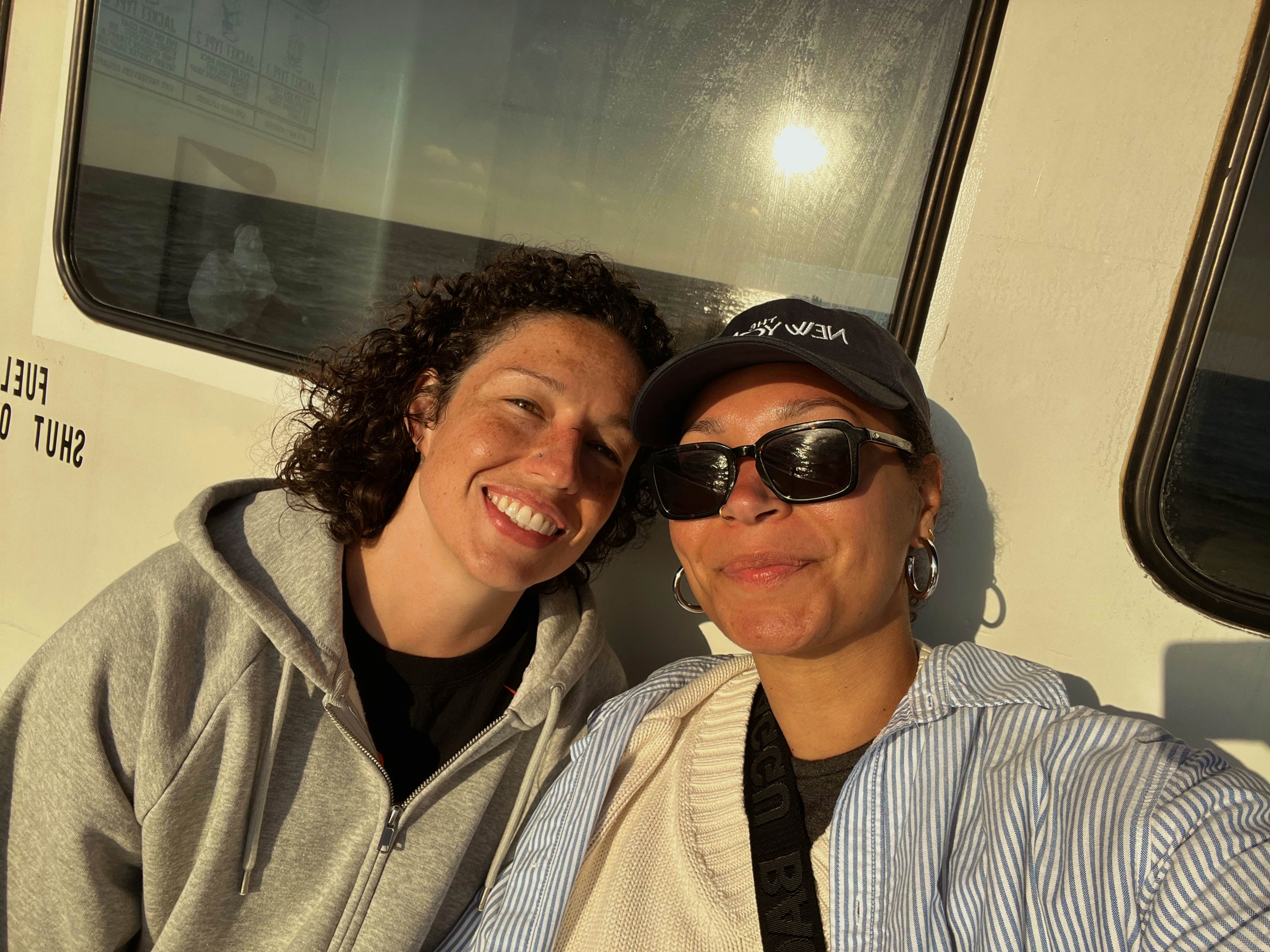 A selfie of two women smiling while sitting on a ferry on a sunny day. One woman is wearing sunglasses and a hat.