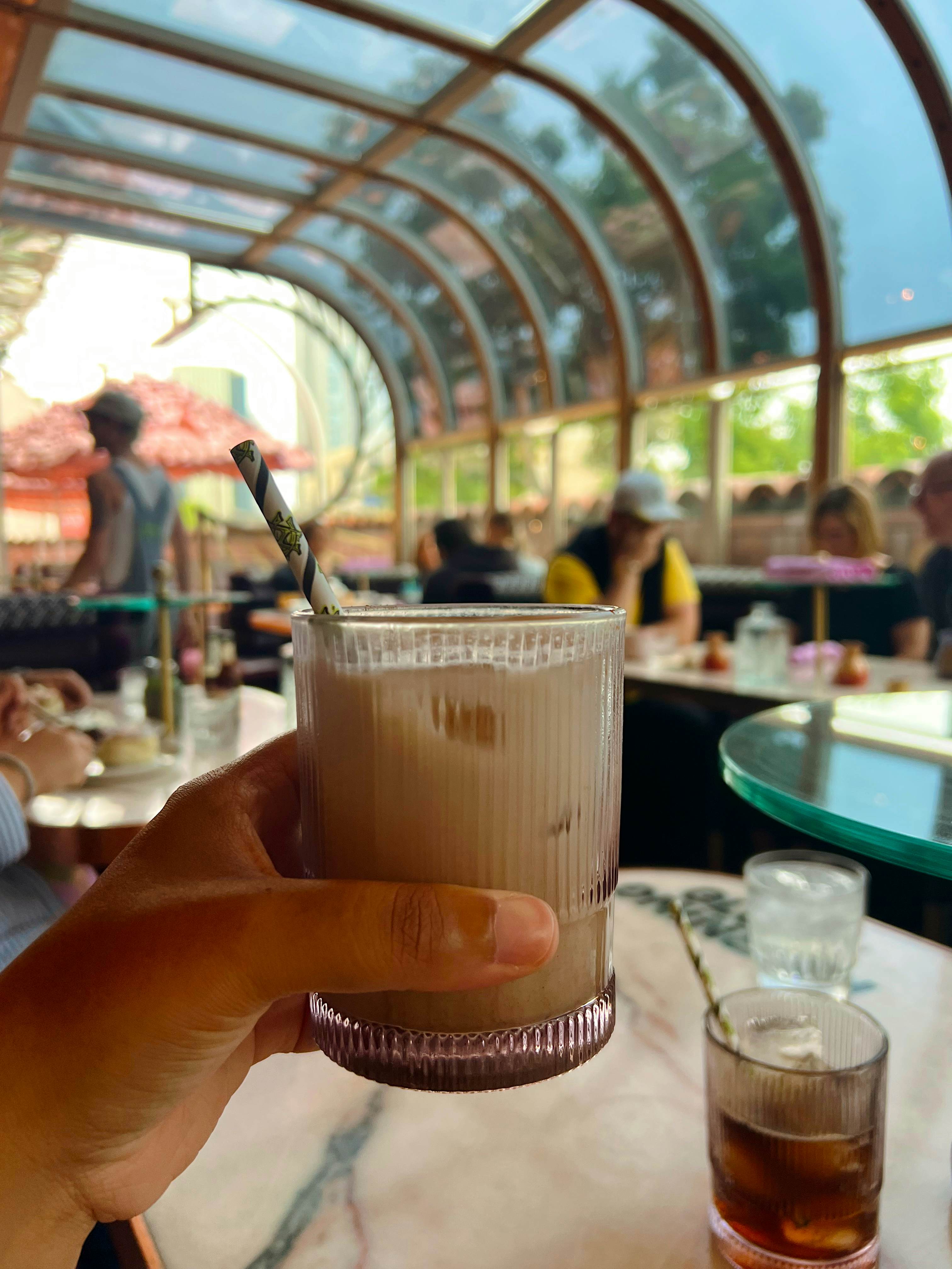 A hand holding a glass cup of iced coffee with a straw with out-of-focus patrons at tables in the background.
