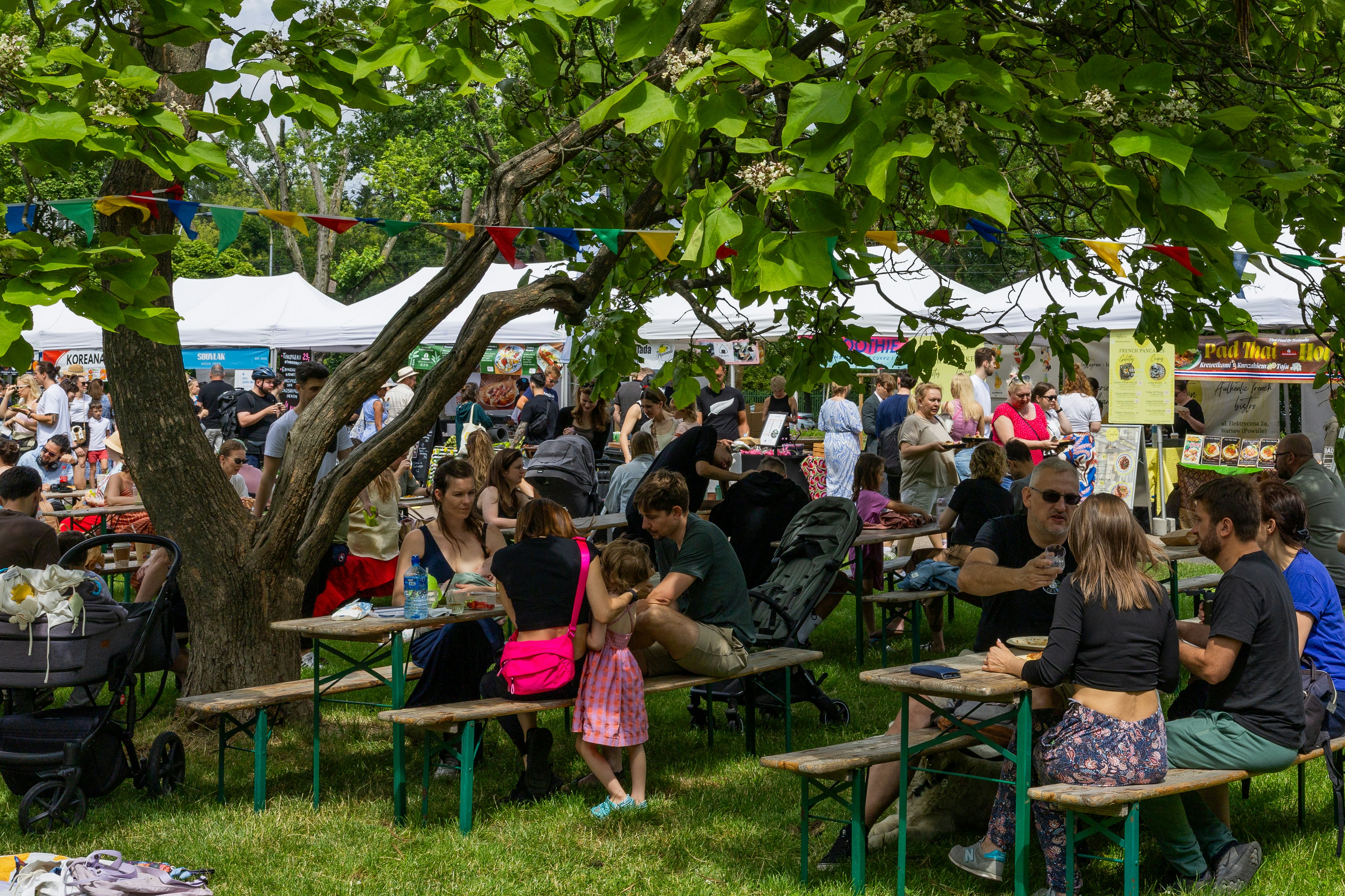 People sit at picnic tables on grass under a tree in Warsaw, Poland; white tents with vendors selling food are in the background.