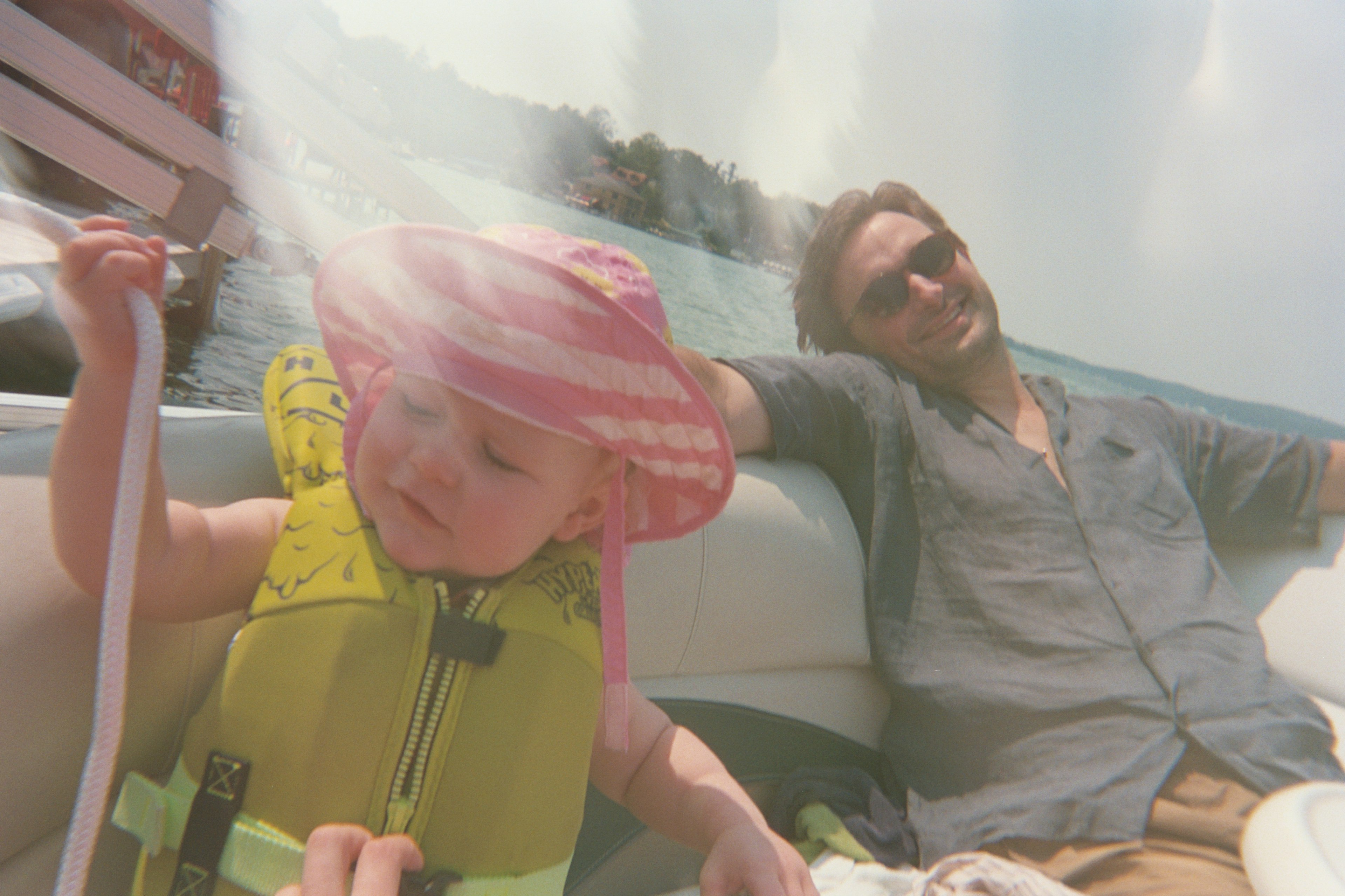 A baby in a pink striped hat and green life vest, a man in a blue shirt lounges in the background