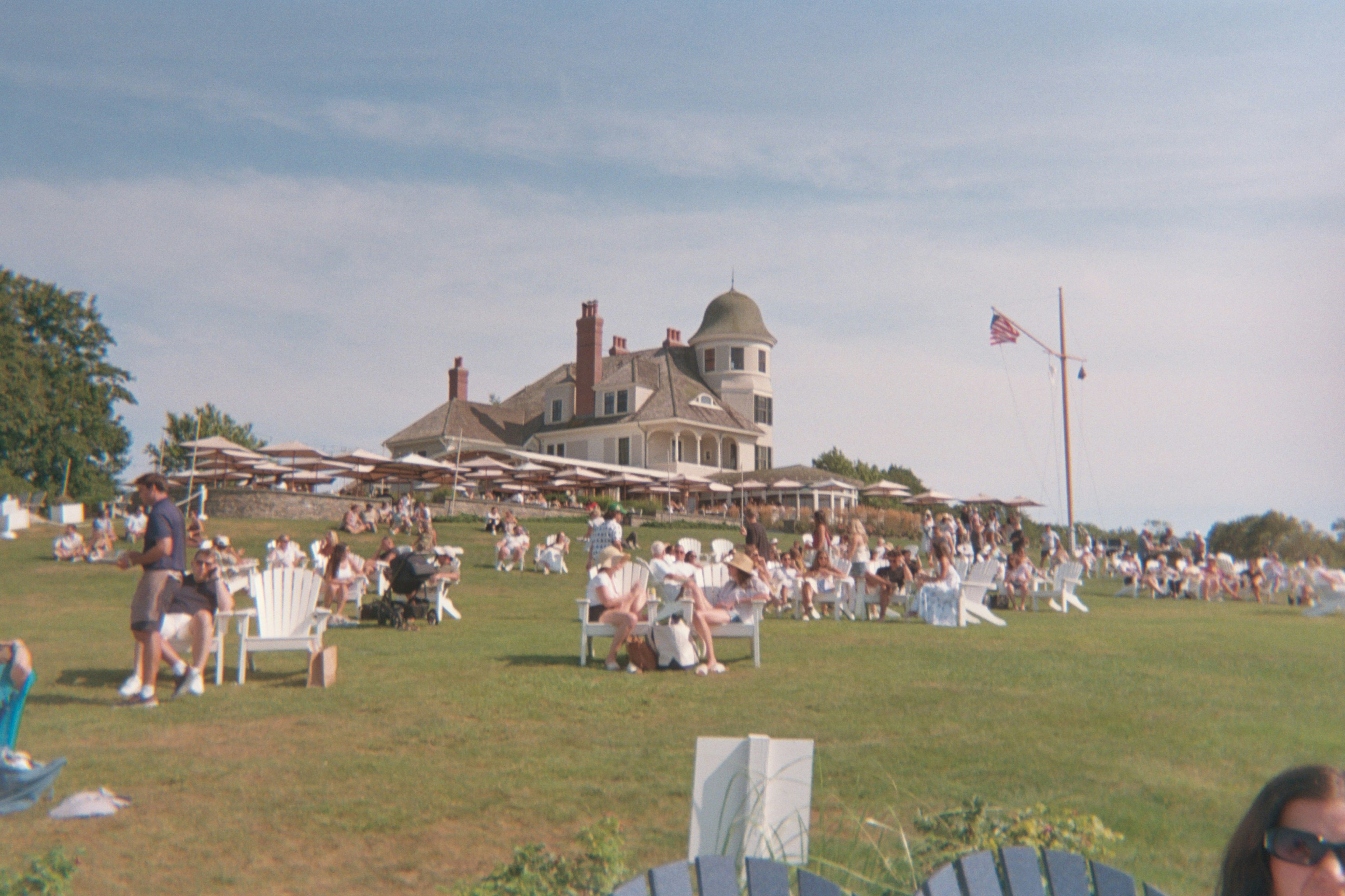 People in Adirondack chairs on a lawn with a large building behind them and an American flag flying overhead