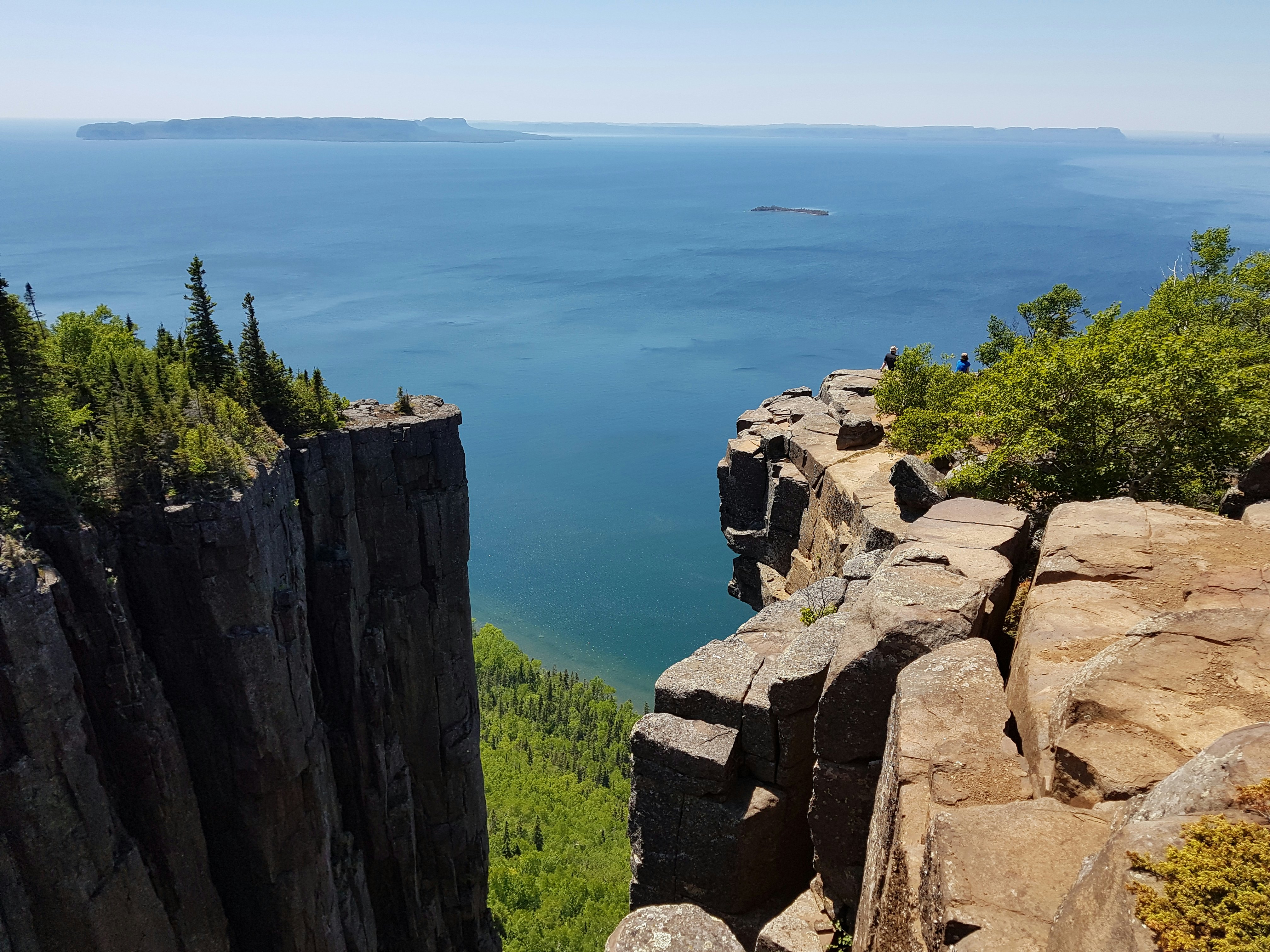 Two tall rock cliffs, separated by a narrow gap, tower over the forested shoreline next to a glassy blue lake on a clear day in Ontario, Canada.