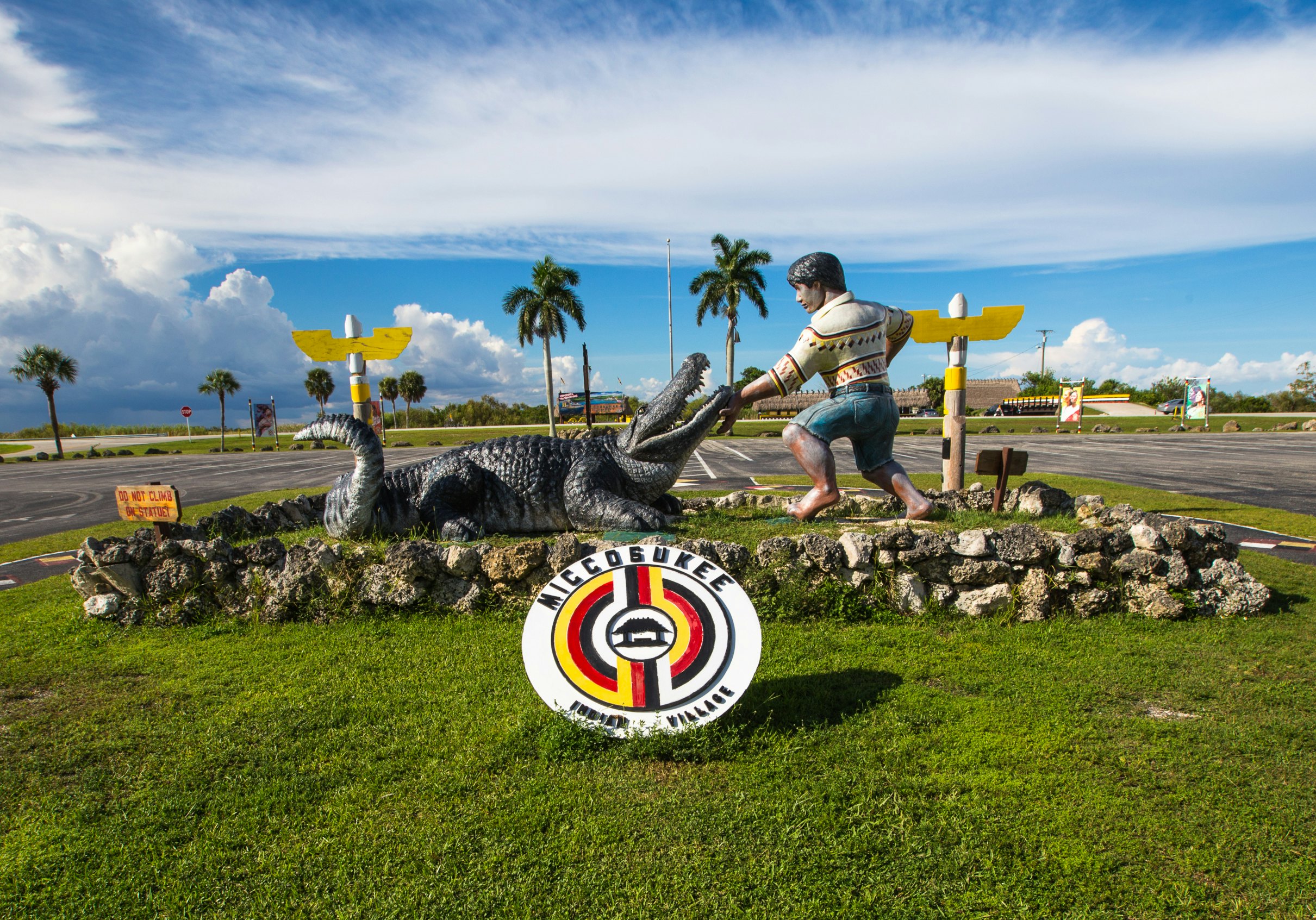 A sculpture of a person with a gator and a large sign welcoming visitors to Miccosukee Indian Village.