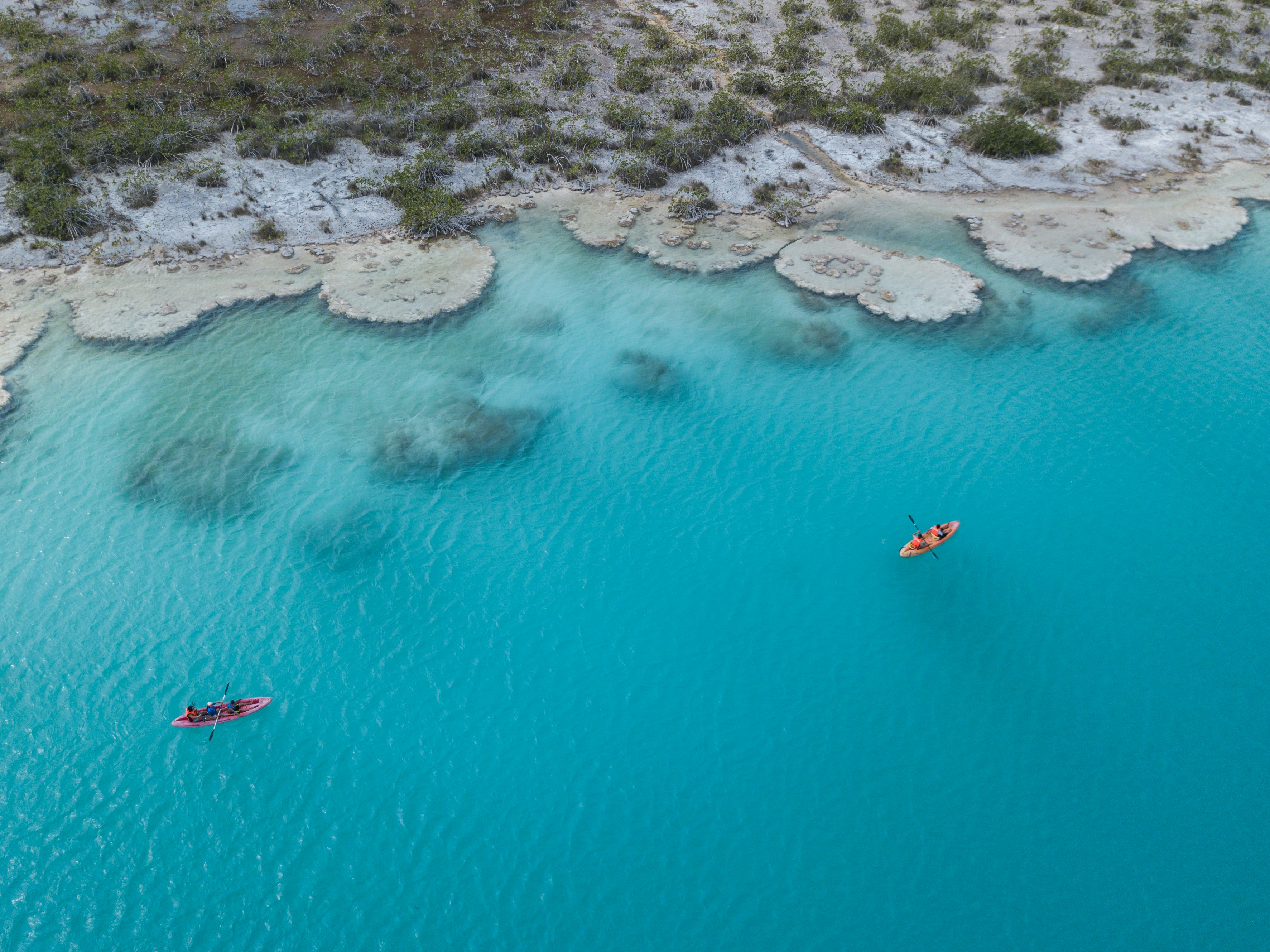 Two kayaks photographed from above gliding through pristine blue waters along a sandy and brush-filled shoreline.