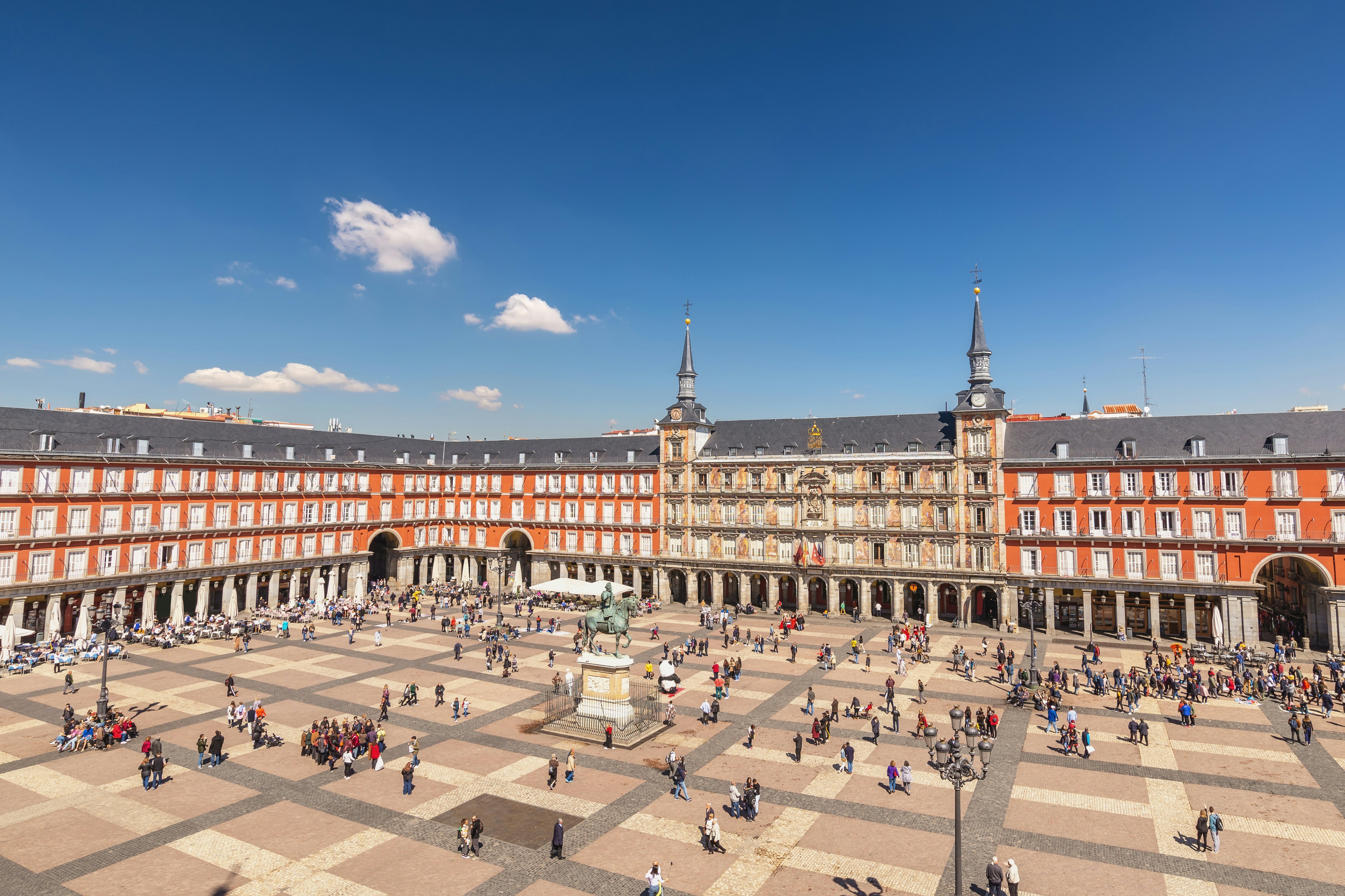 A busy square surrounded by an expansive building. The square is full of people with a statue of a person on a horse in the middle.
