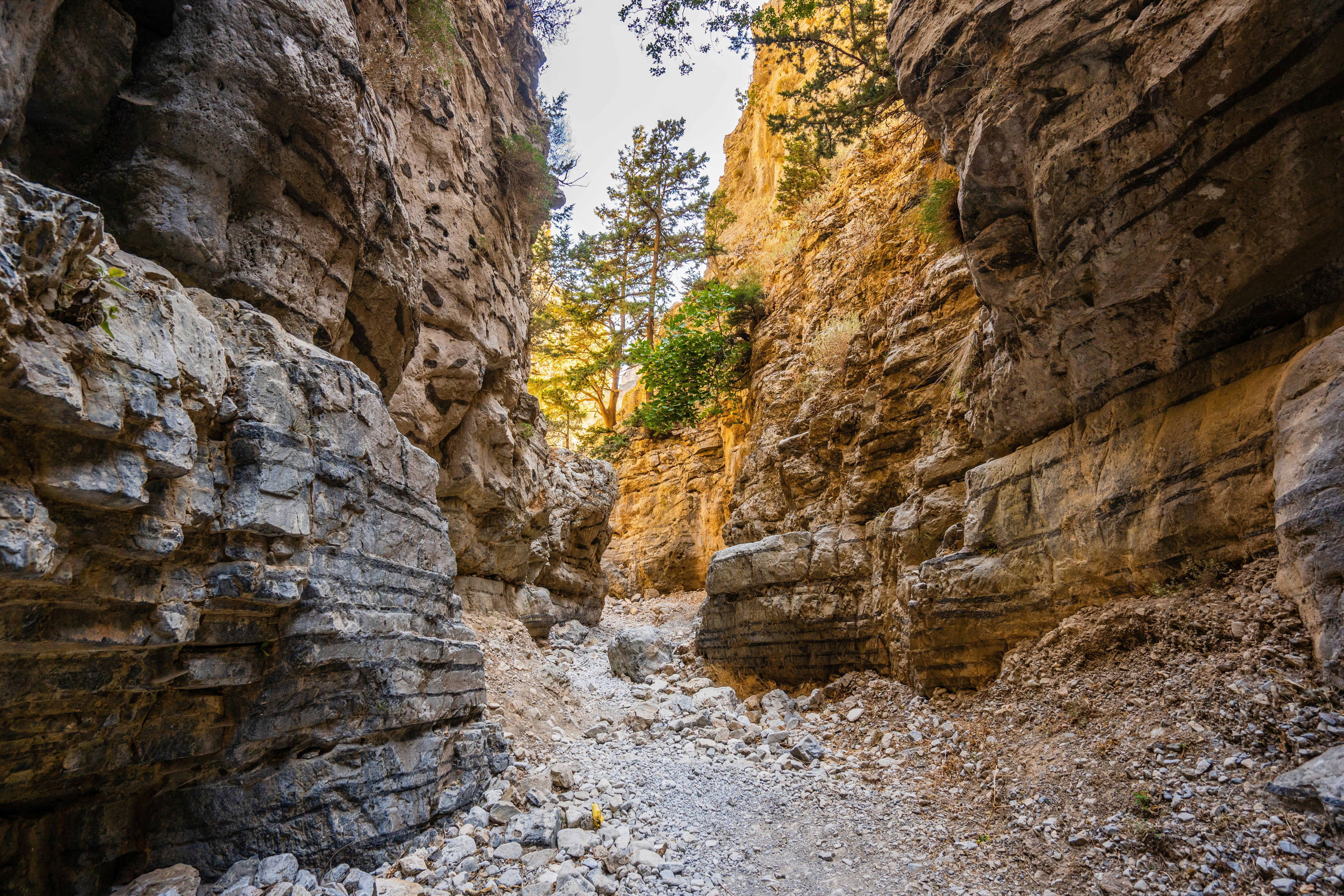 A rocky path through a gorge in Crete; the rock walls are high, and there is sunlight shining on the rock face at the opening at the end.