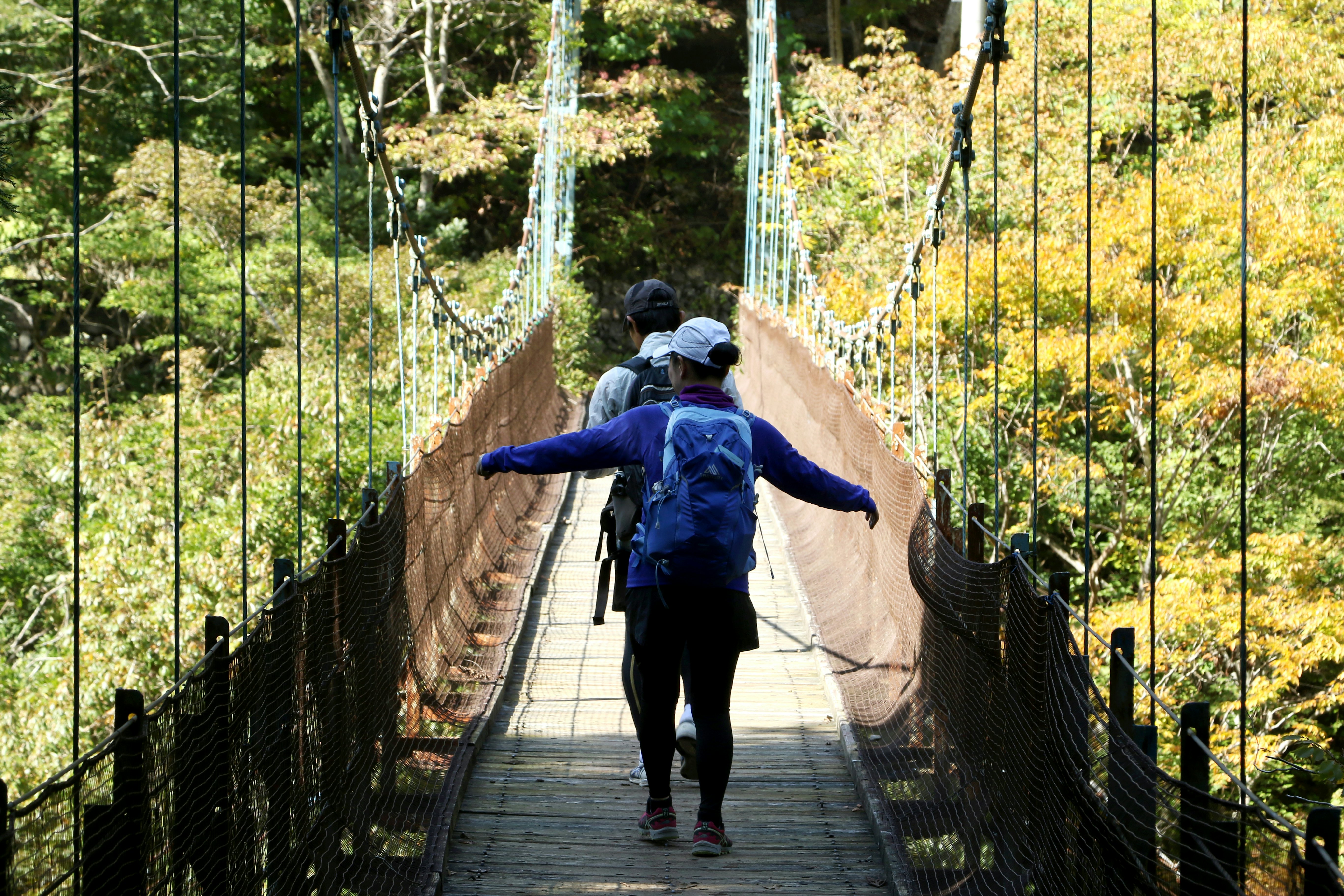 Two people with backpacks and wearing ballcaps walk single file on a suspension bridge on a sunny day; the person walking in back has their arms out to the side.