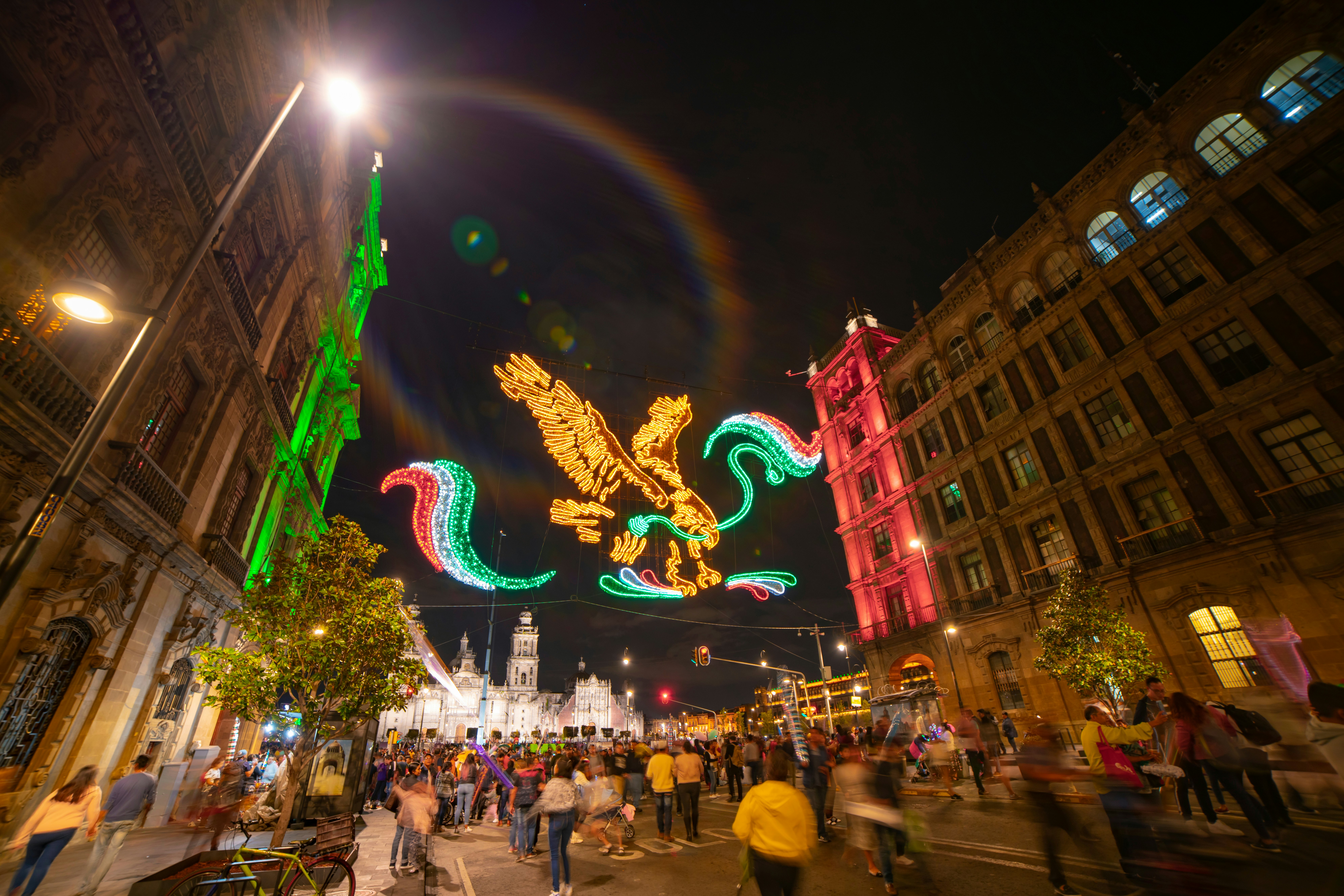 People walk on a city street at night. Illuminated decorations hang over the street, and a cathedral is illuminated in the distance.