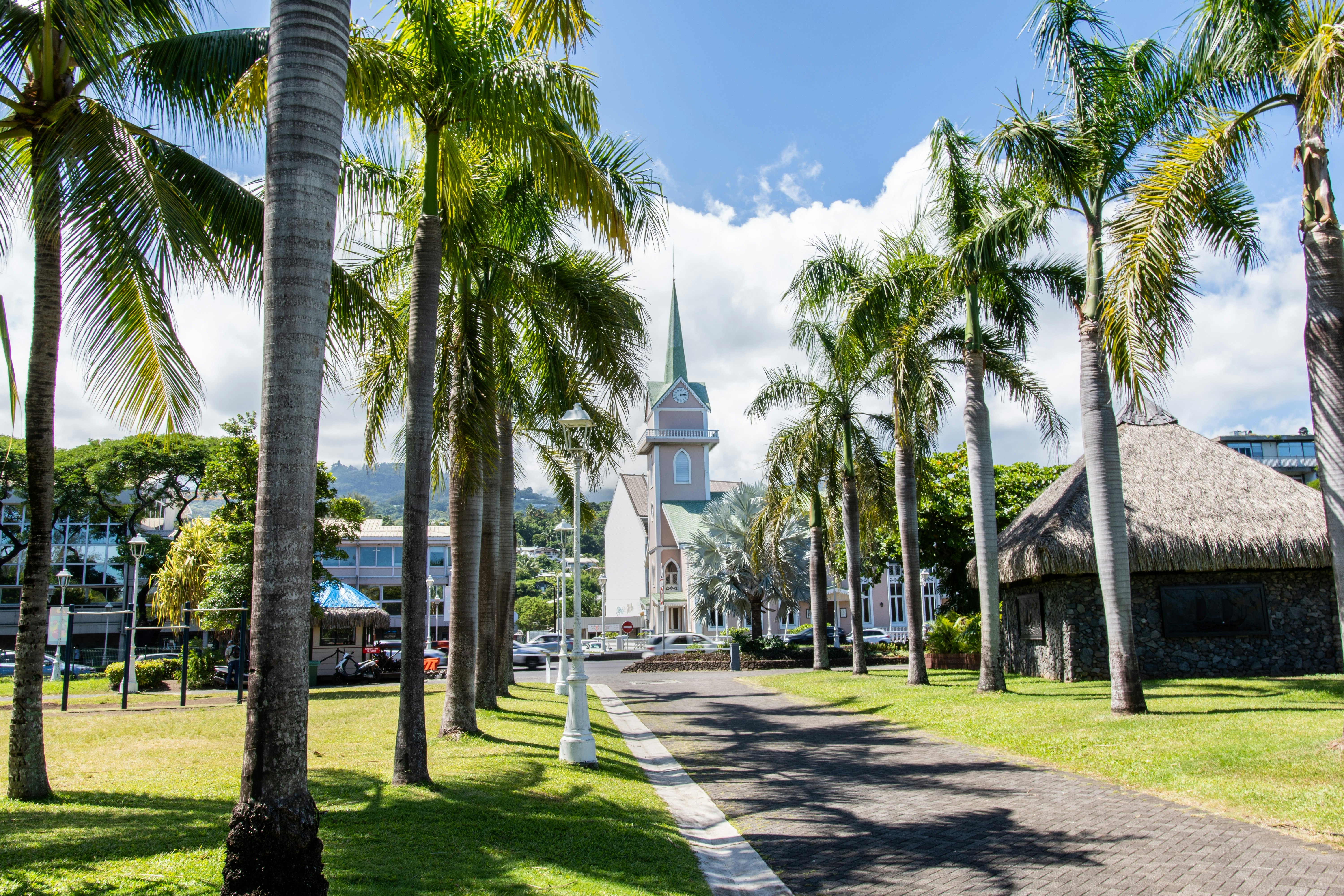 A pal tree-lined path through a garden leads towards a grey and white church with a clock and pointed steeple.