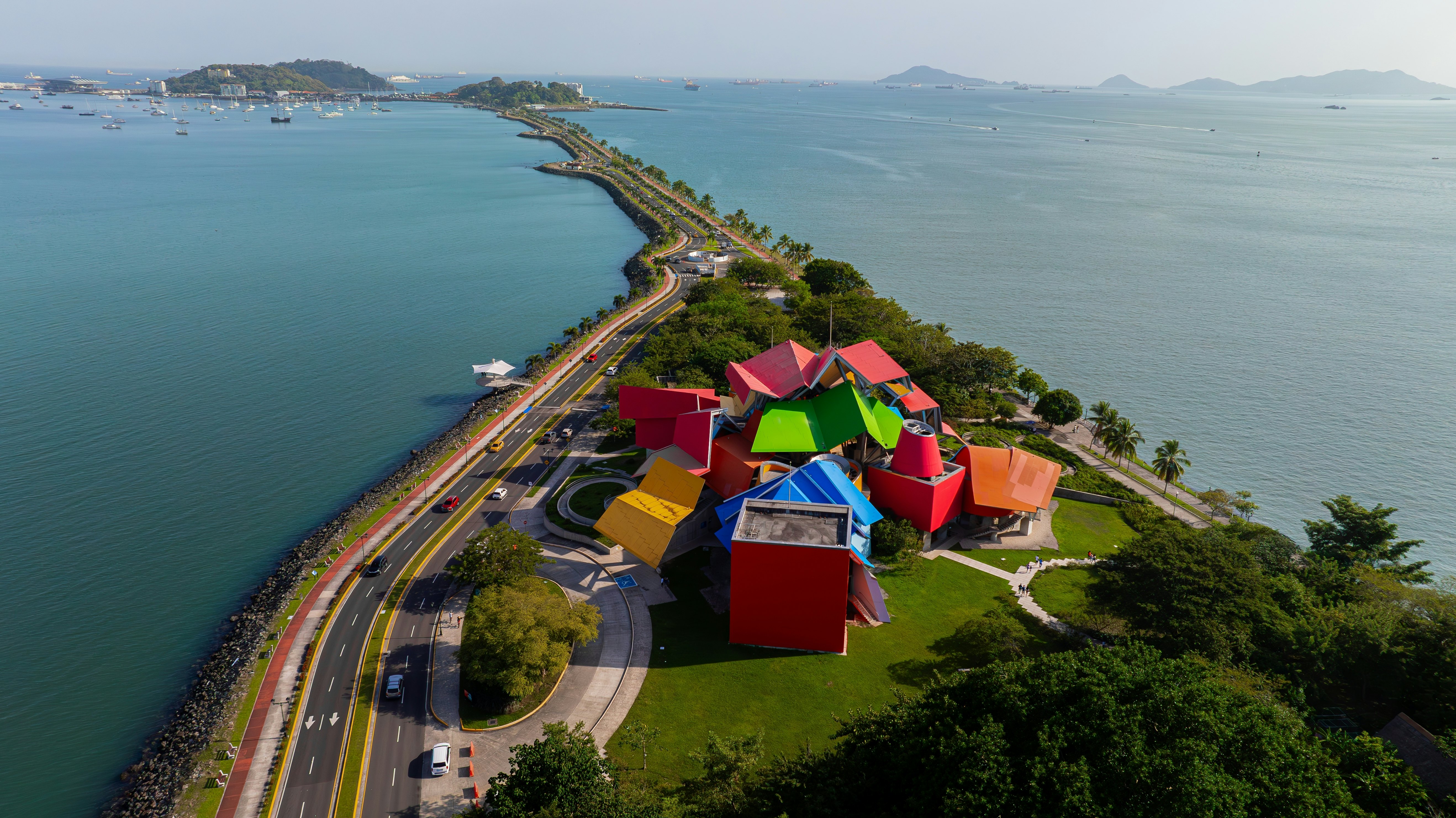 A building with rainbow-colored roofs on a lush, green land with water on both sides and a paved roadway on the left side of the photo and a walkway on the right side.