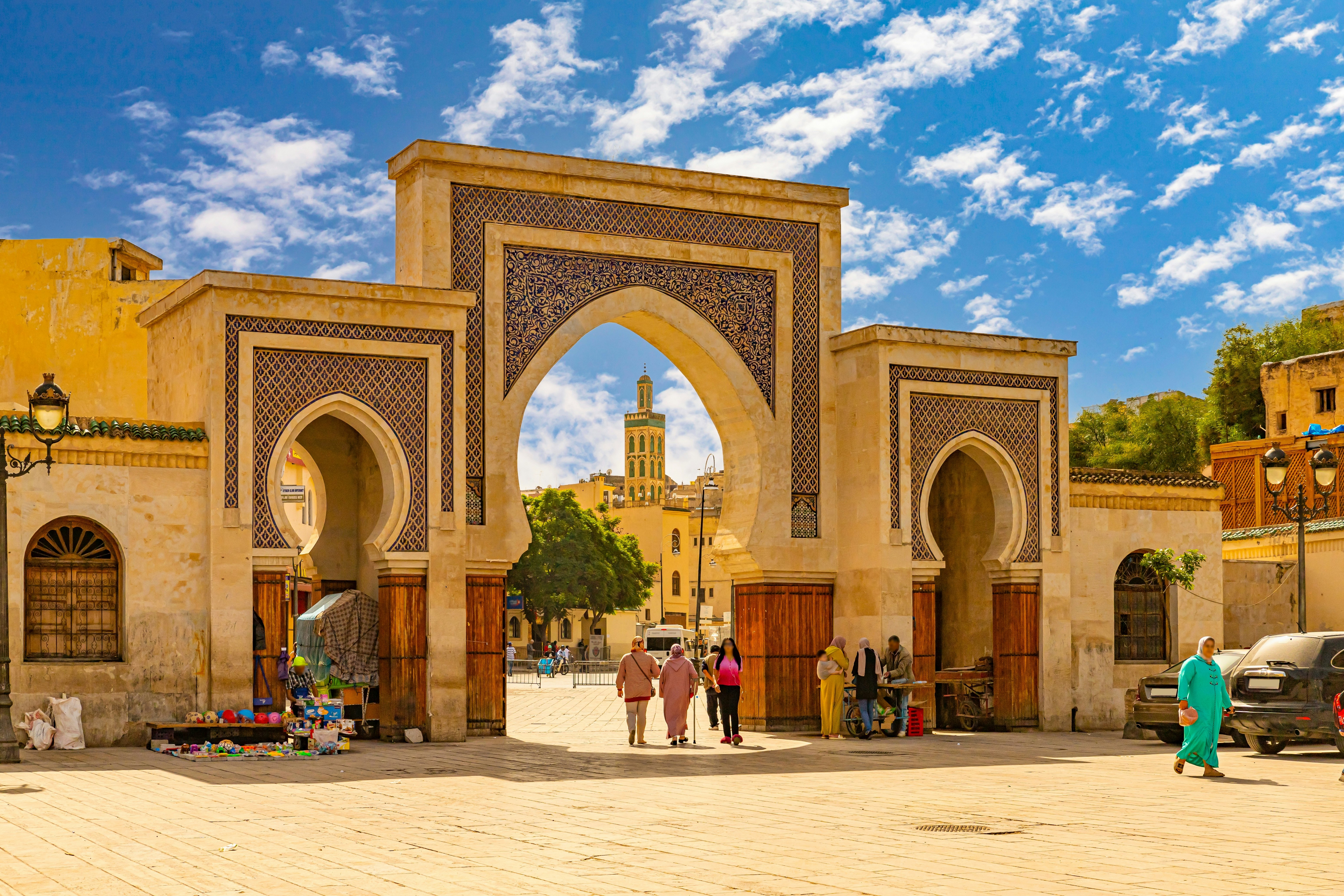 People walk through a historic gateway into an old-town center with a tall mosque tower.