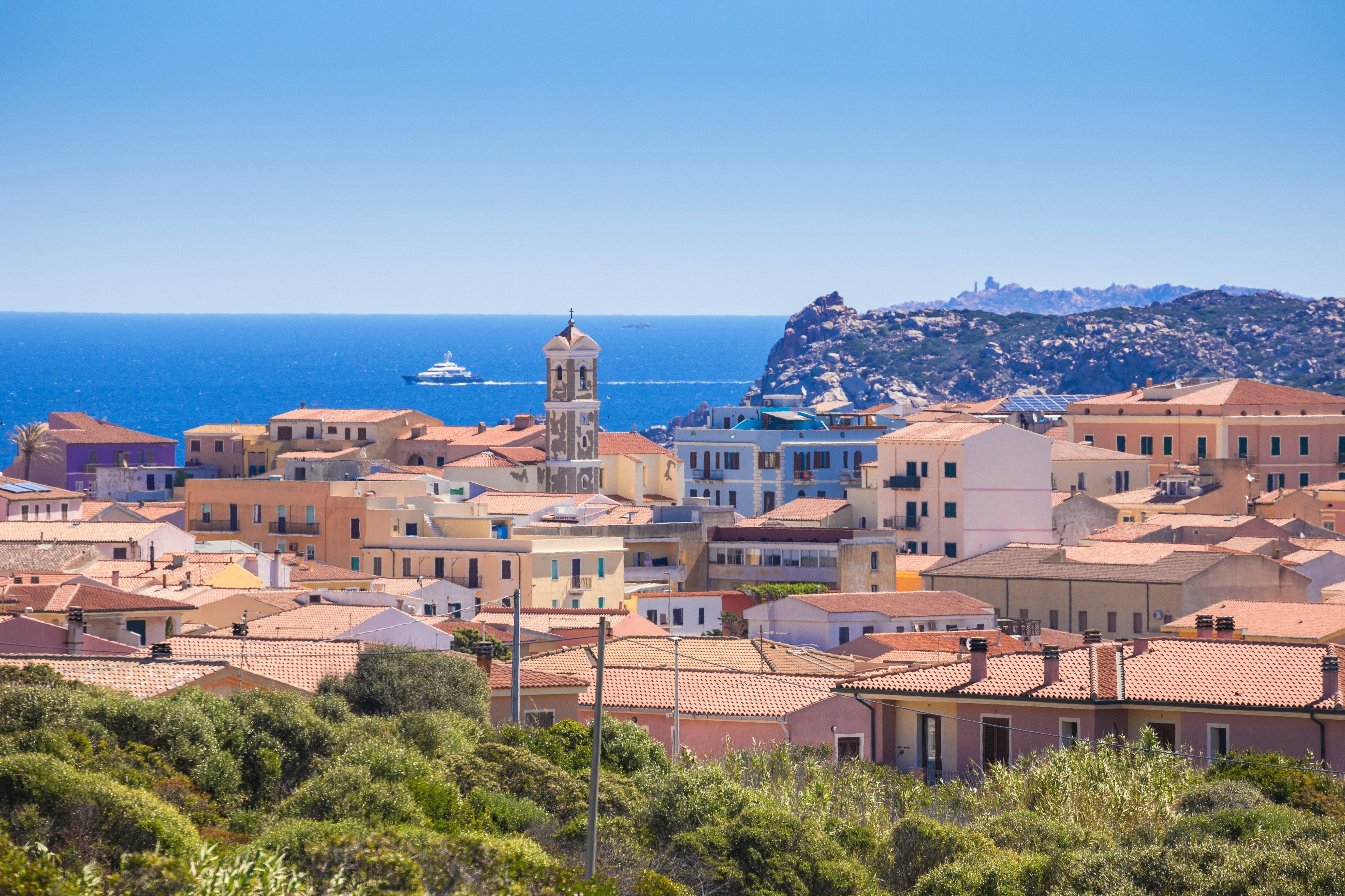 A town in Sardinia set against the blue sea; there are terra cotta roofs on low buildings and a tall tower in the center.