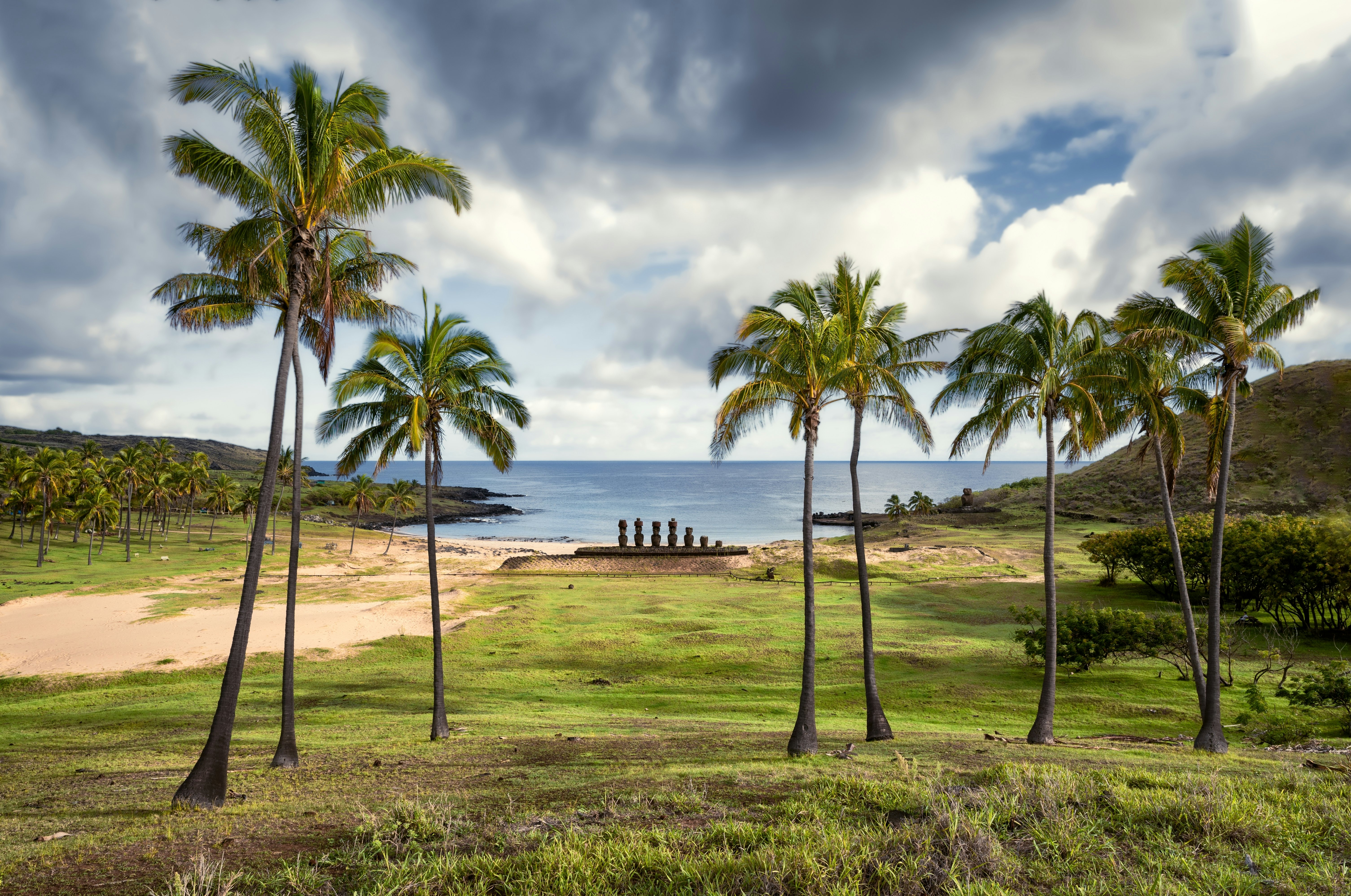 View of Anakena beach with palm trees and moai statue silhouettes during a sunny day, with dramatic cloudy sky, Easter Island, Chile.