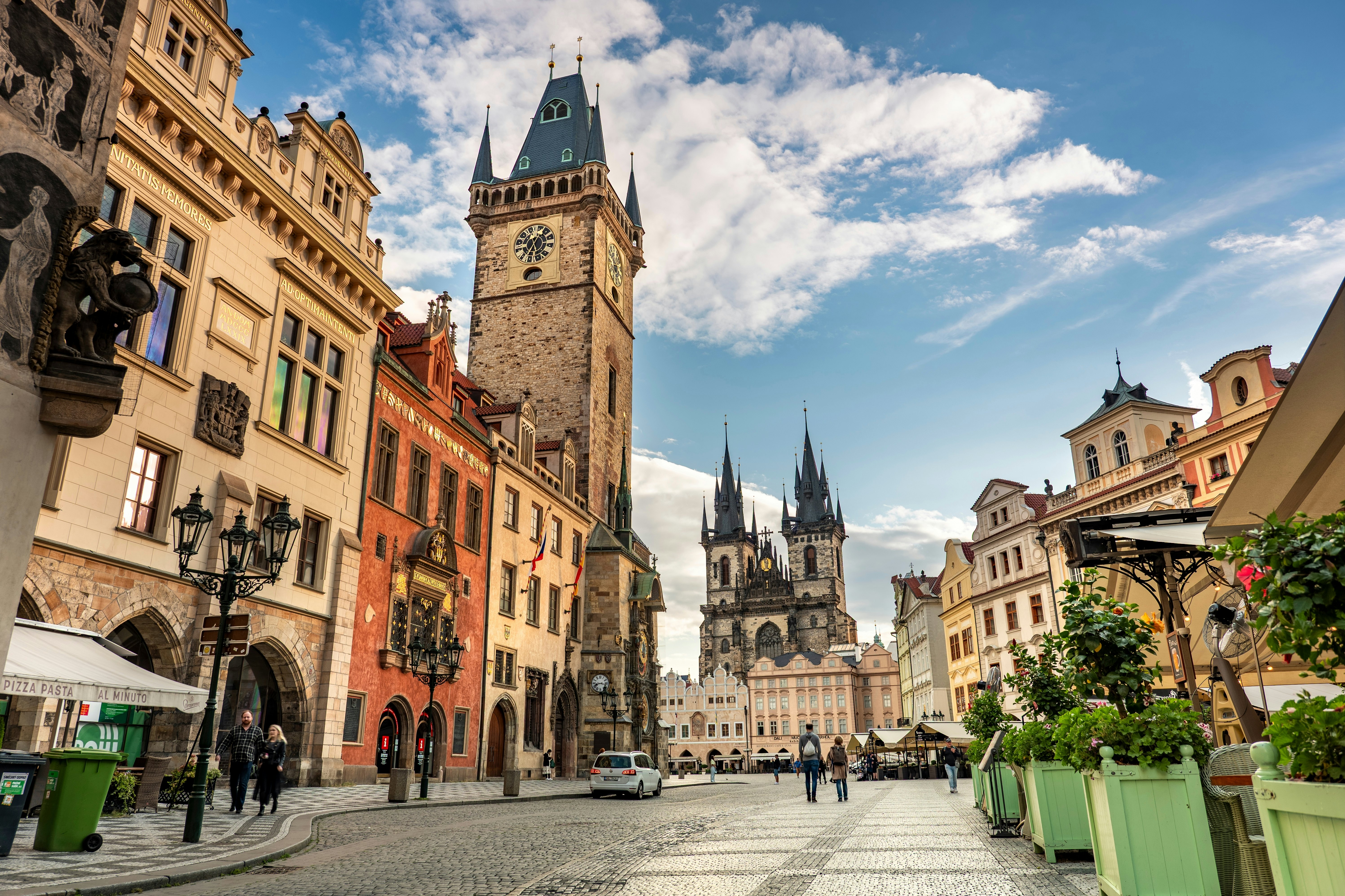 A medieval astronomical clock tower rises over a square in Prague; the spires of a cathedral are at the far end of the square, and other historic buildings surround the open space.