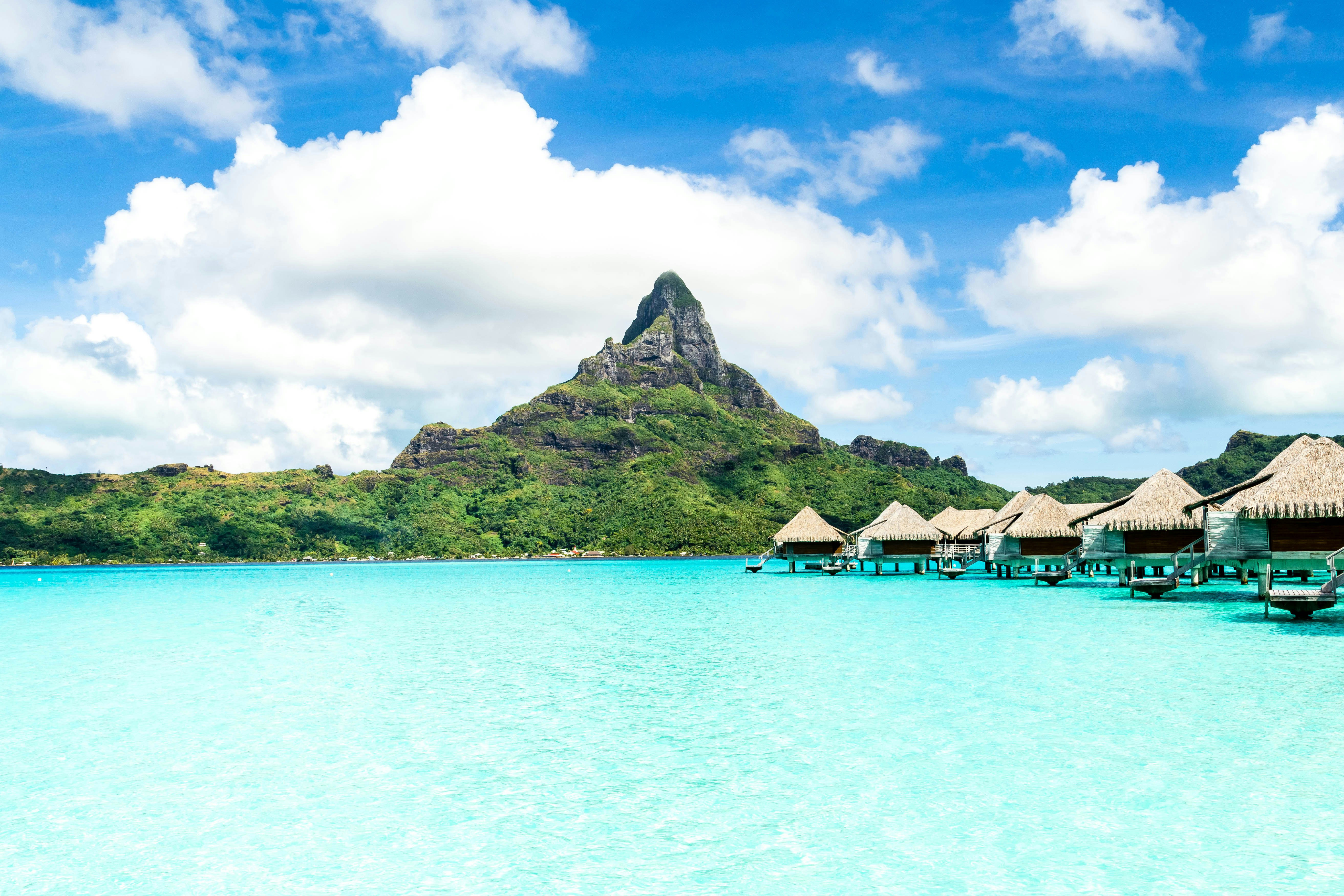 Clear ocean water laps at the edge of wooden bungalows built over the sea. A pointed mountain peak rises above on the nearby island.