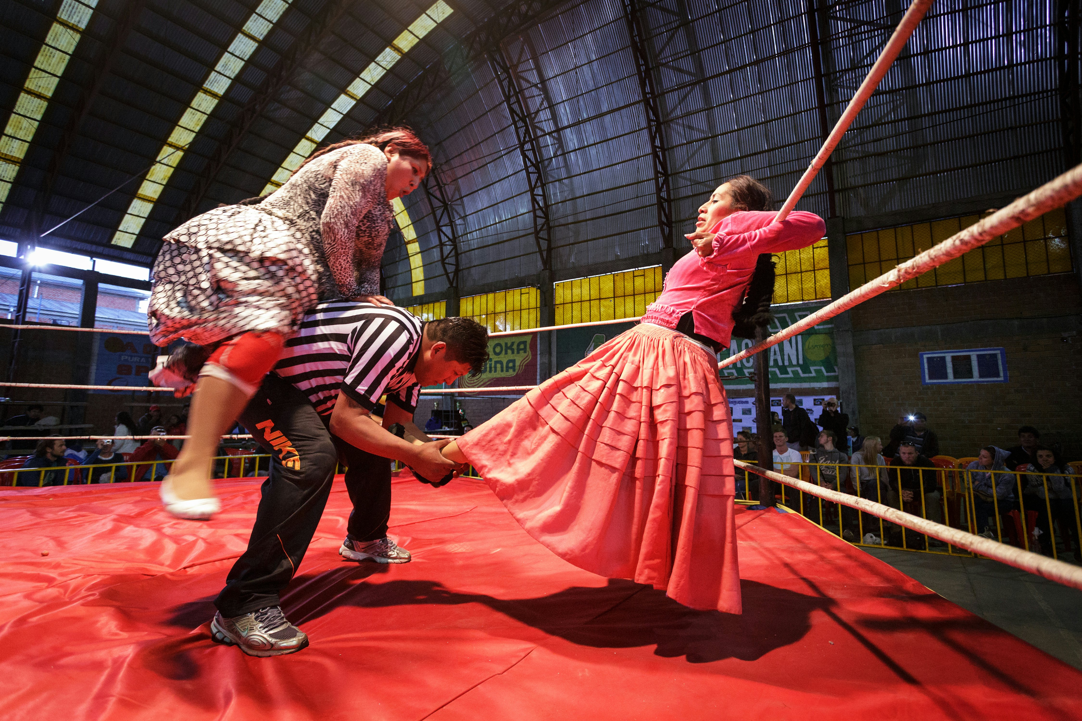 A woman in a wrestling ring leans on the ropes as her ankles are grabbed by the referee while another woman leaps on his back.
