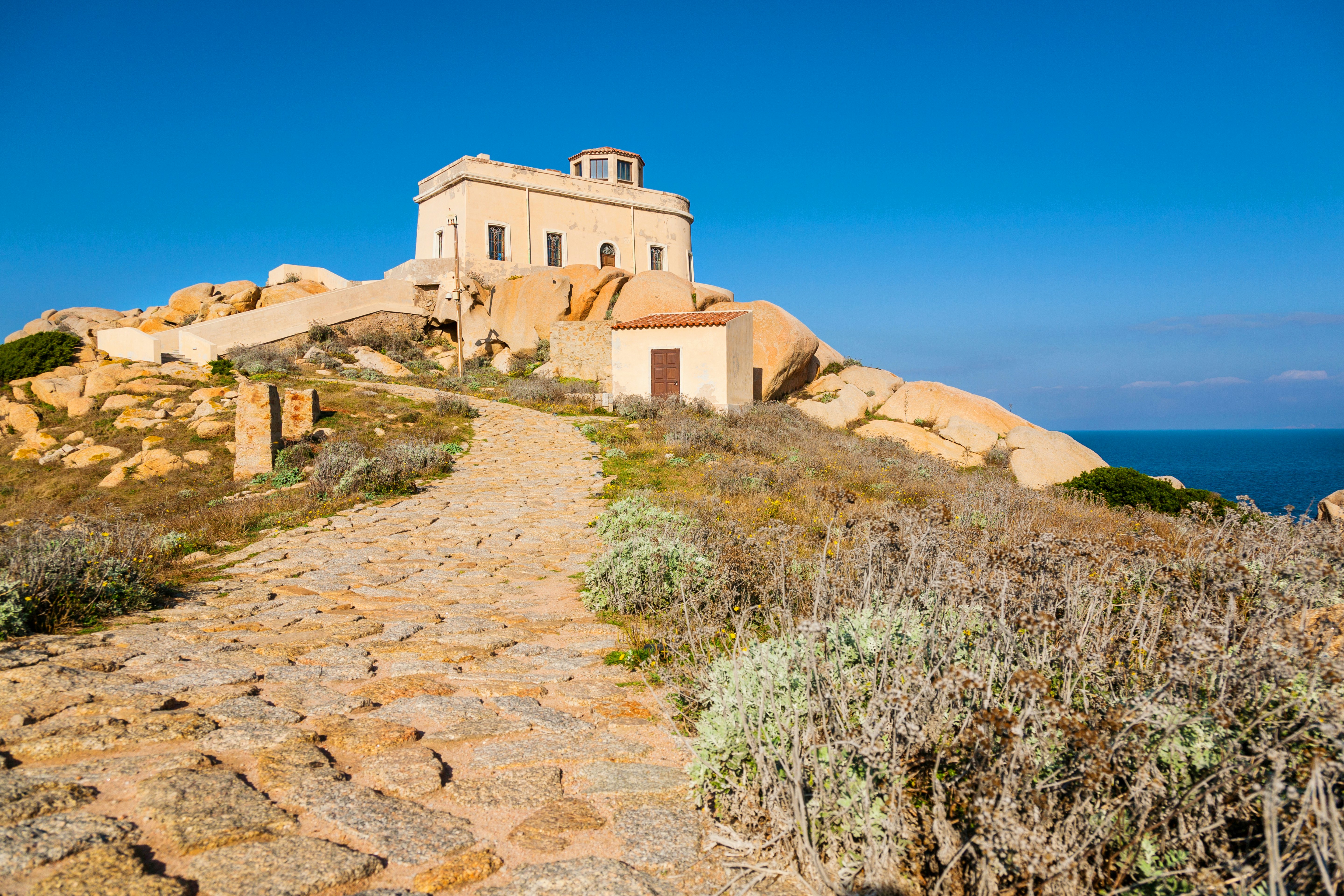 A stone pathway with scrub vegetation to the side leads to a golden-colored building with a small lighthouse at the top.