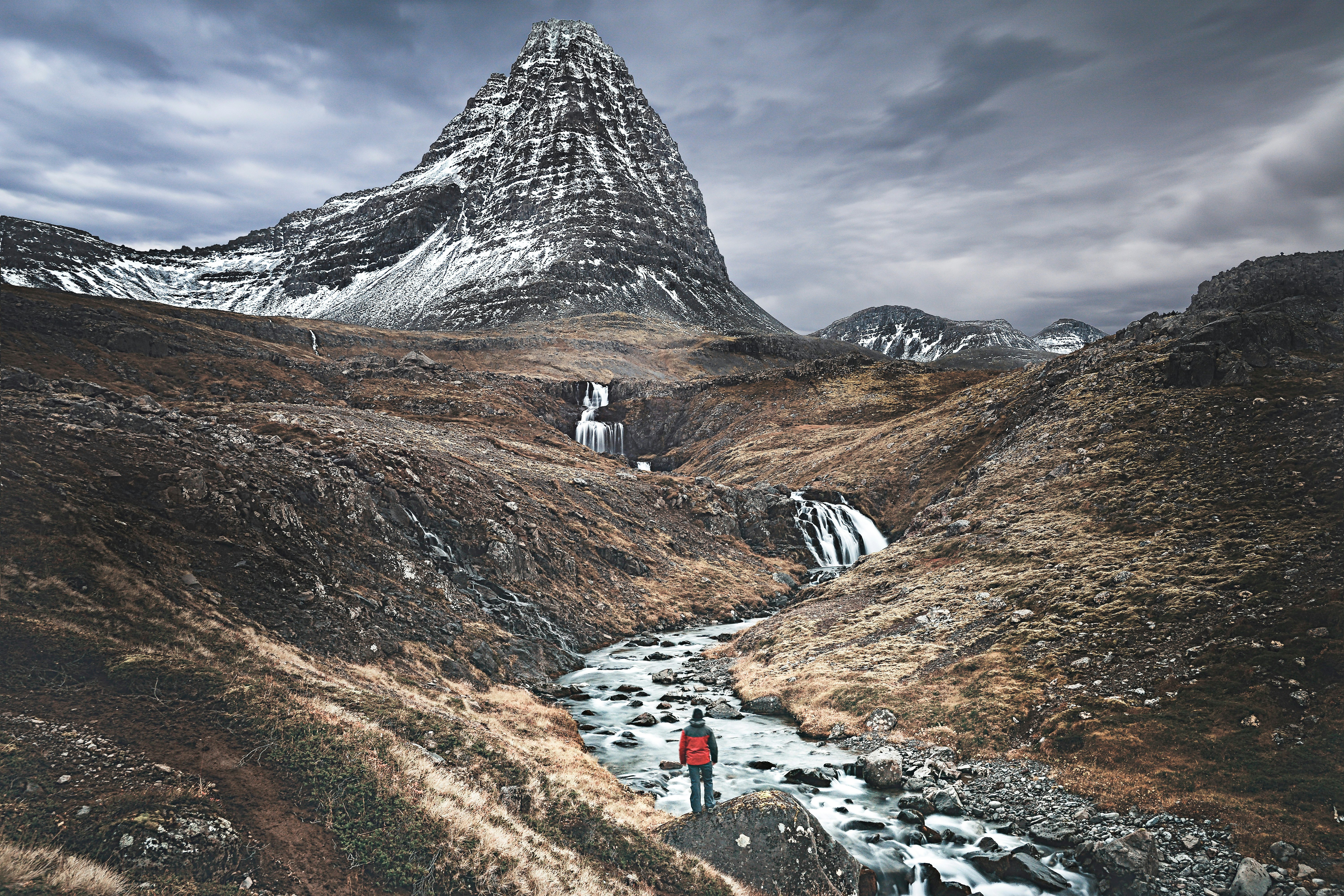 A lone hiker, seen from behind, stands on a rock overlooking a stream with waterfalls and snow-dusted mountains in the distance.