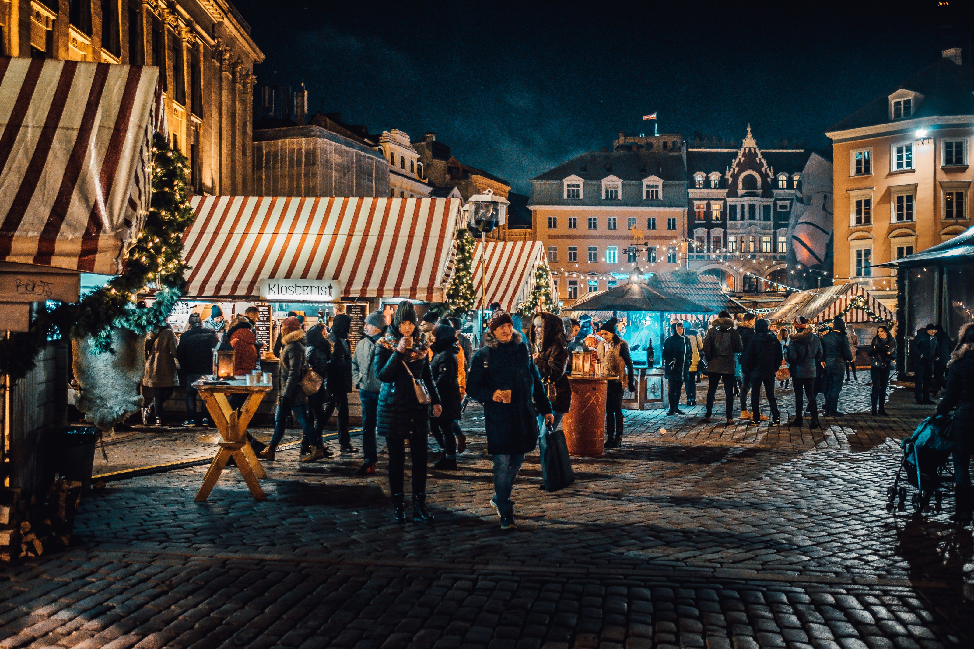 People gather at a Christmas market in a cobbled square in Riga at night. People queue for hot drinks at a red and white striped stand and carry shopping bags.